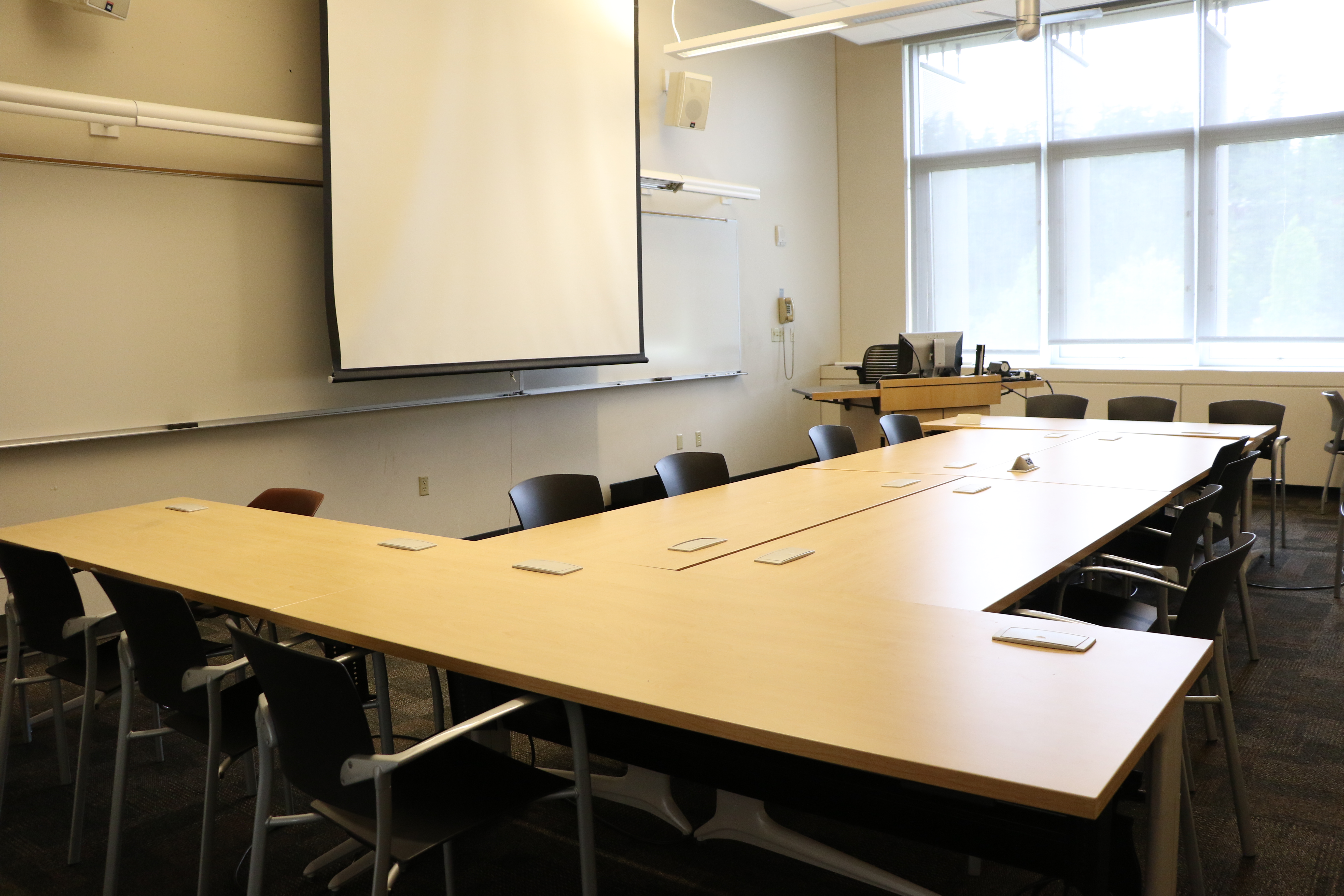 Room consists of carpet, moveable tables and chairs and a podium and whiteboard located at the front of the room. 