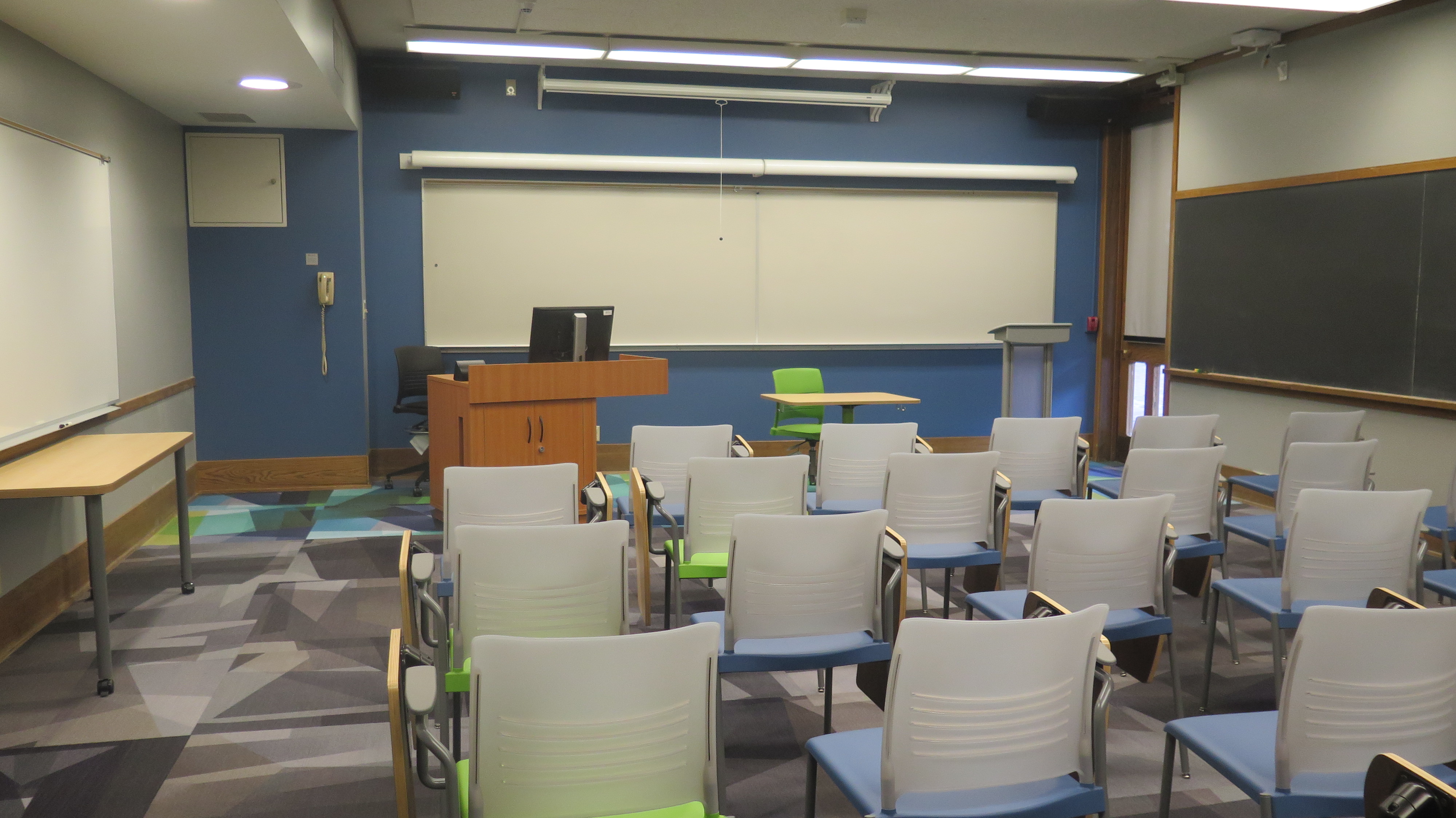 View from the back of the room, Moveable tablet arm chairs, Carpet floors, White board on the front wall, chalkboards on the outer walls of classroom.