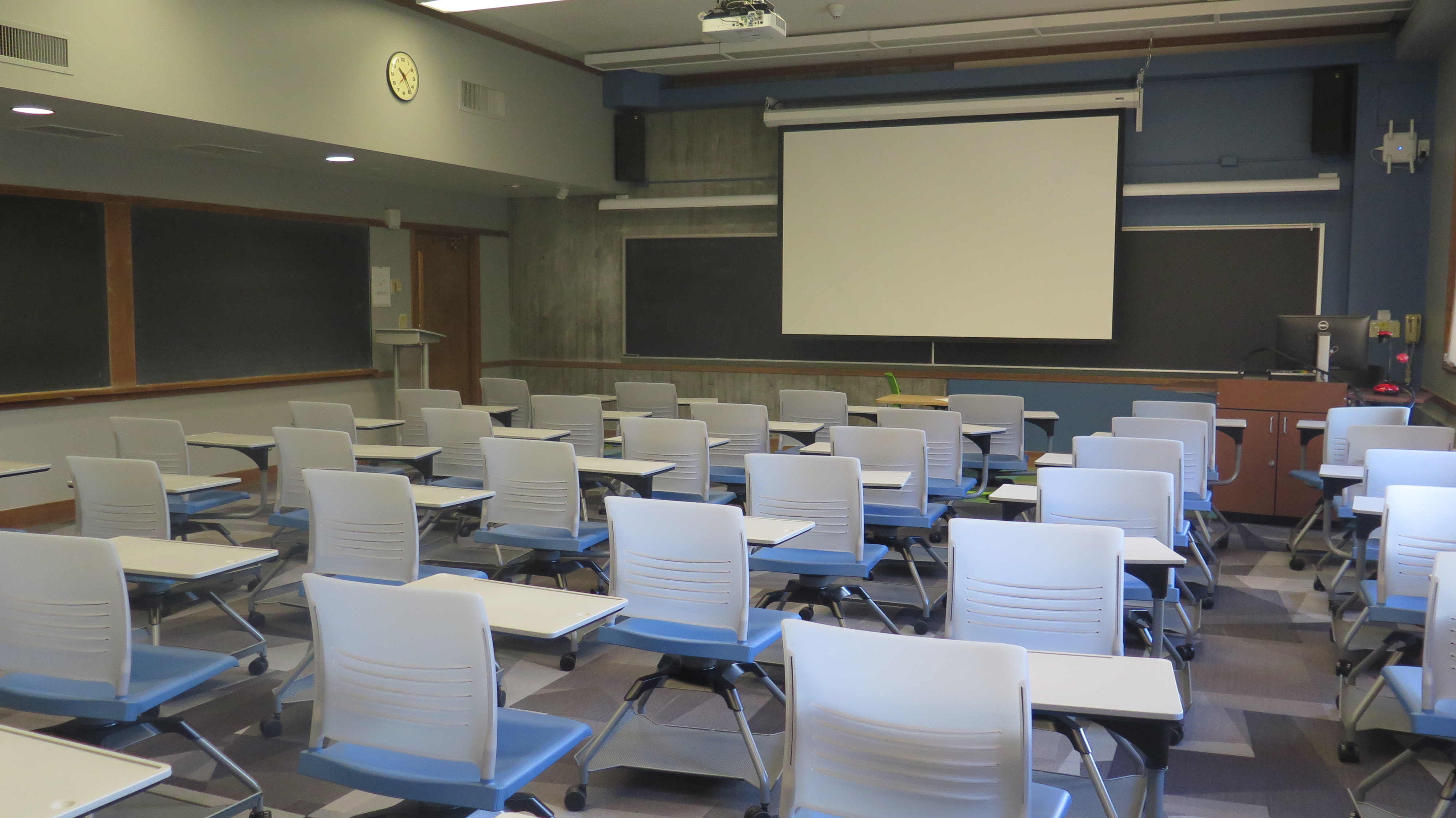View of Bond Hall 225 from the back of the room, Podium at the front of room. carpet floor, movable tablet arm chairs, chalkboard at the front of class, whiteboard spread out on the side walls of classroom