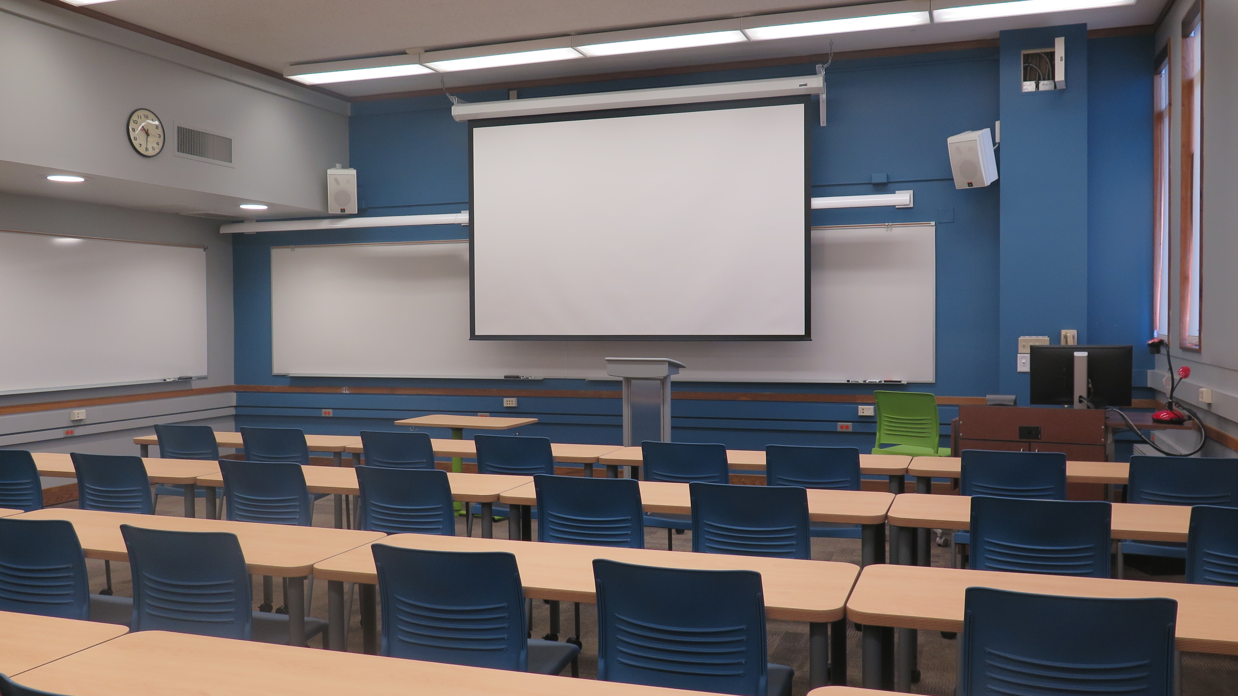 View of Bond Hall 317 from the back of the room, Movable tables and chairs, podium and whiteboards at the front of the room, whiteboards on one side of the room.