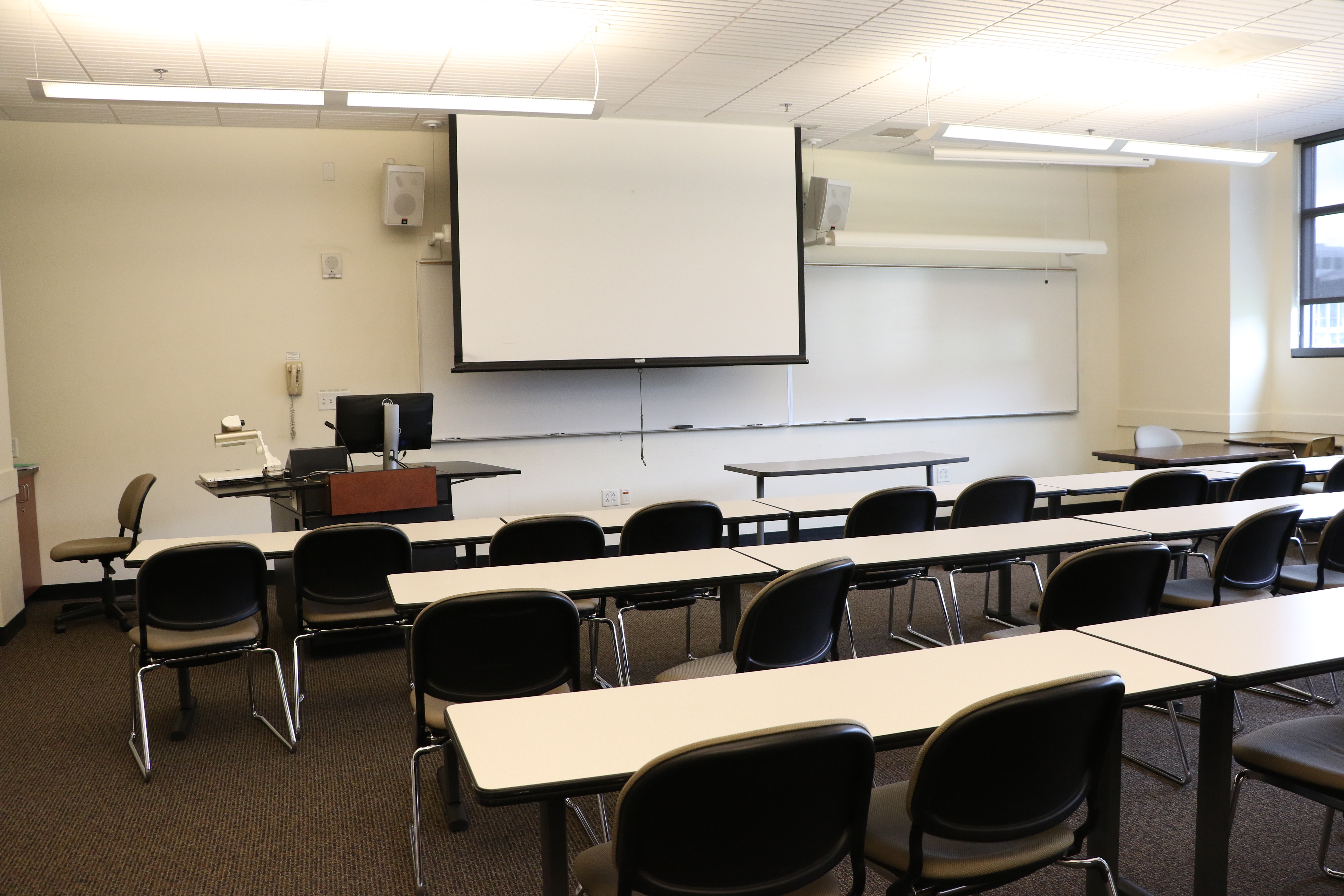 Room consists of carpet, moveable tables and chairs, another table at the front of the room, podium and whiteboard located at the front of the room. 