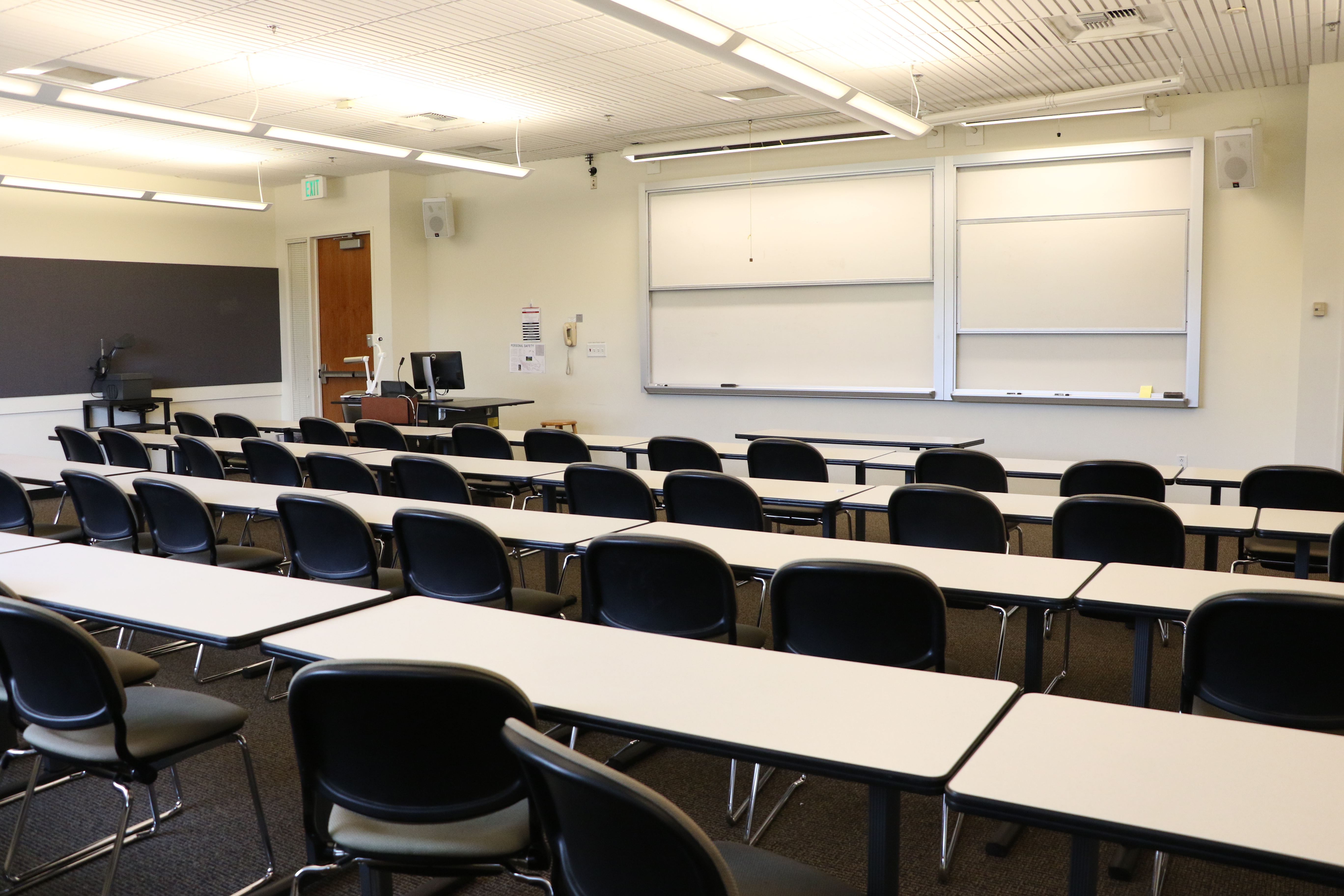 Room Consists of carpet floors, moveable tables and chairs, a white board and podium are both located at the front of the room.