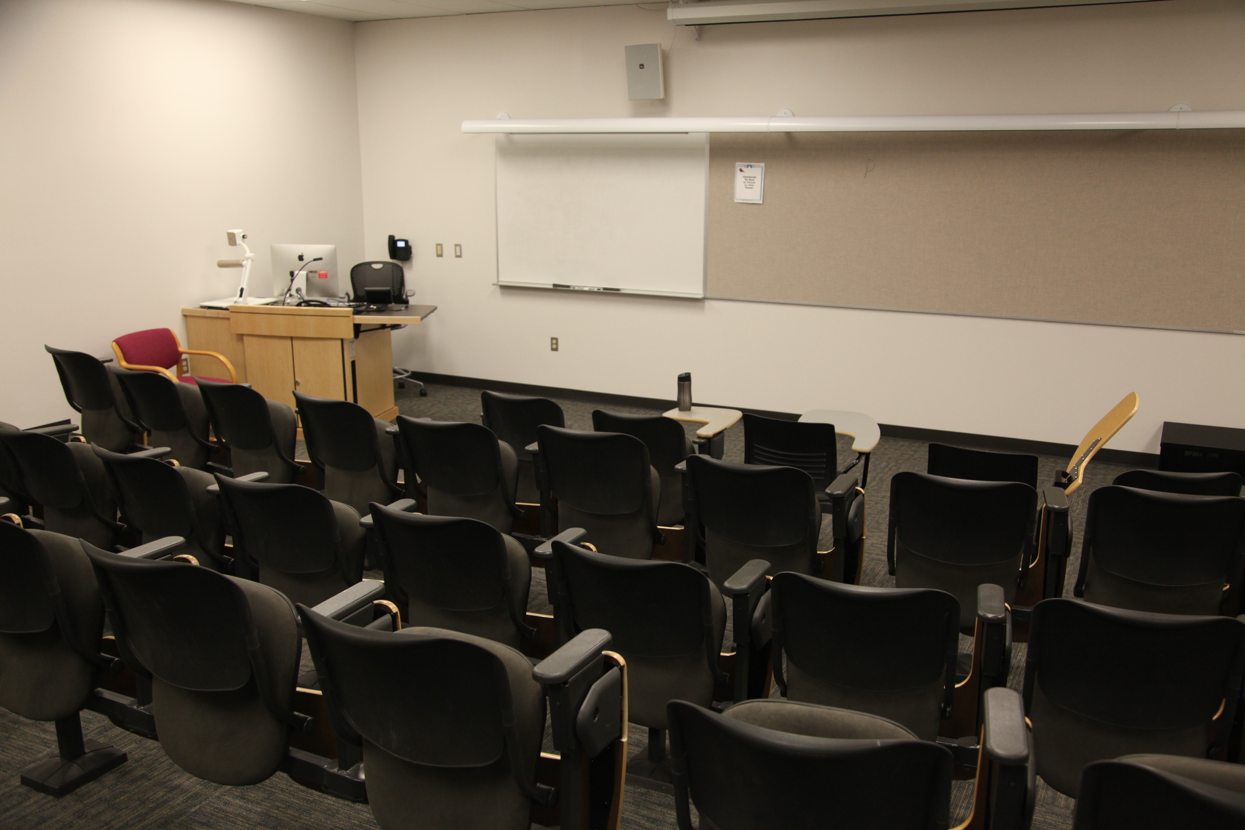 Room Consists of stationary tablet armchairs, White board and podium are located at the front of the room.