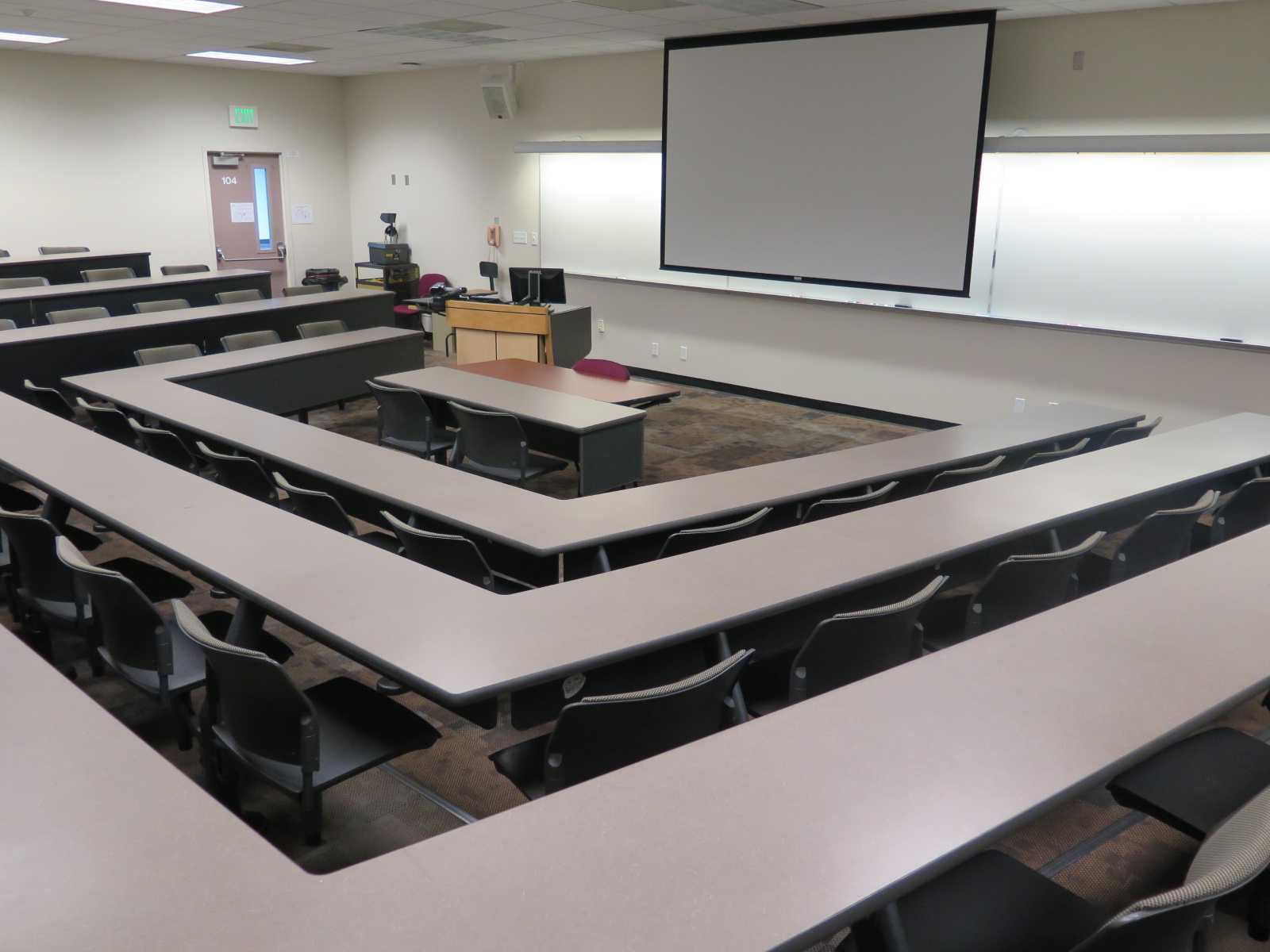 Room Consists of Carpet floors, Stationary rows of Tables and Chairs, and a white board and podium are both located at the front of the room.