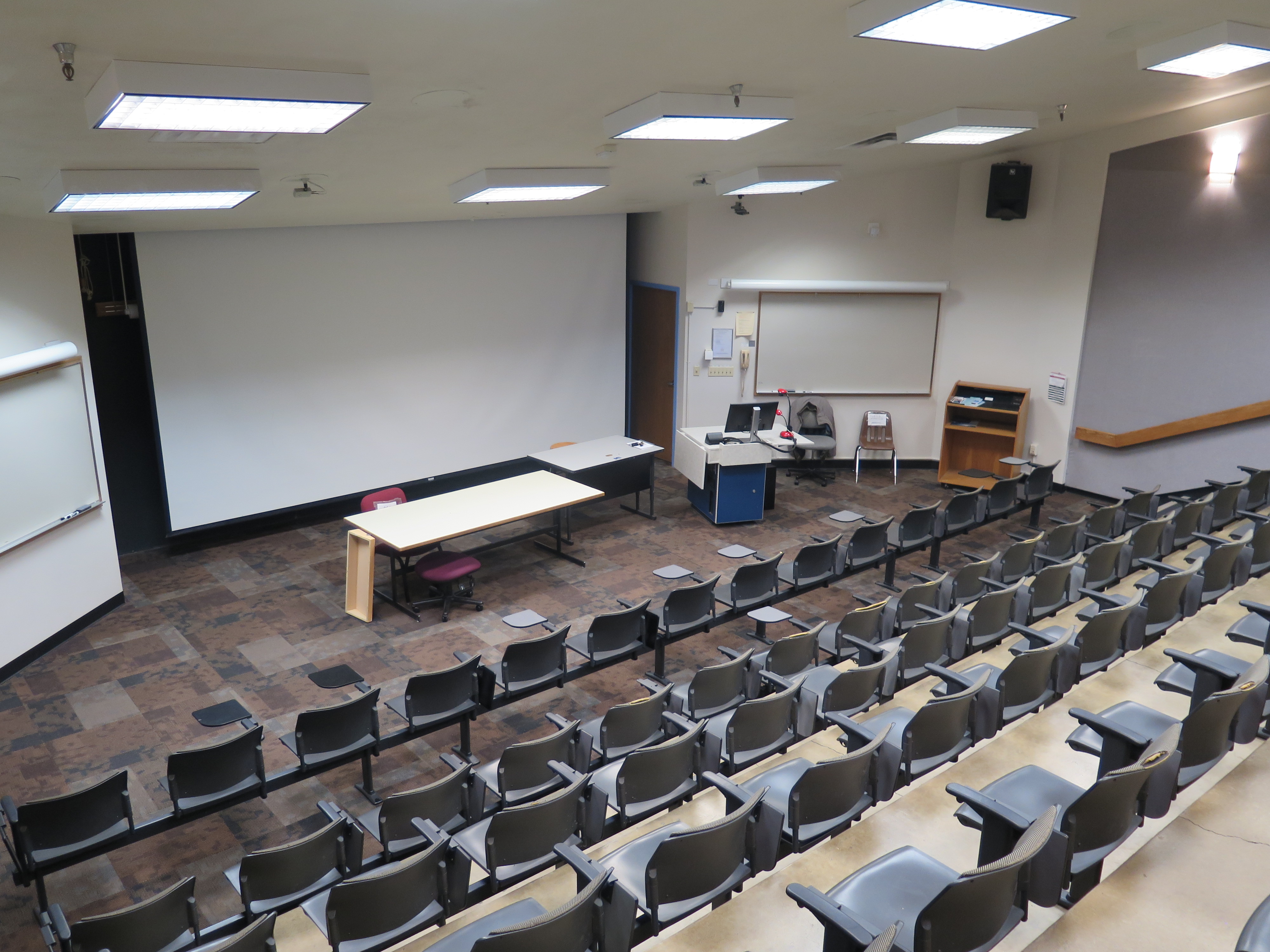 Room Consists of carpet floors, Stationary rows of tablet armchairs in auditorium style with each row of seats going down a level until you reach the front of the room, a white board and podium are both located at the front of the room. 