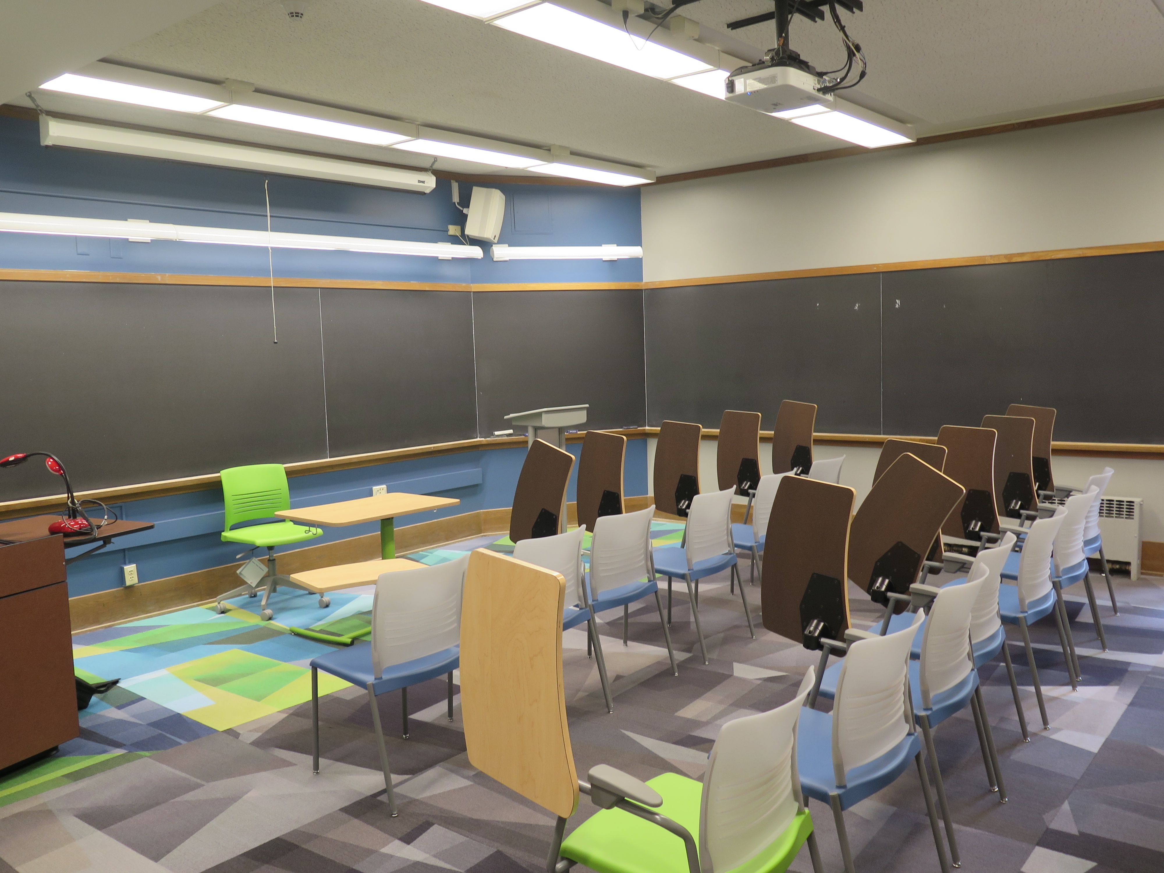 View from the back of the room, Carpet floors, Moveable tablet arm chairs. Podium at the front of room. Chalk boards on three of four walls.