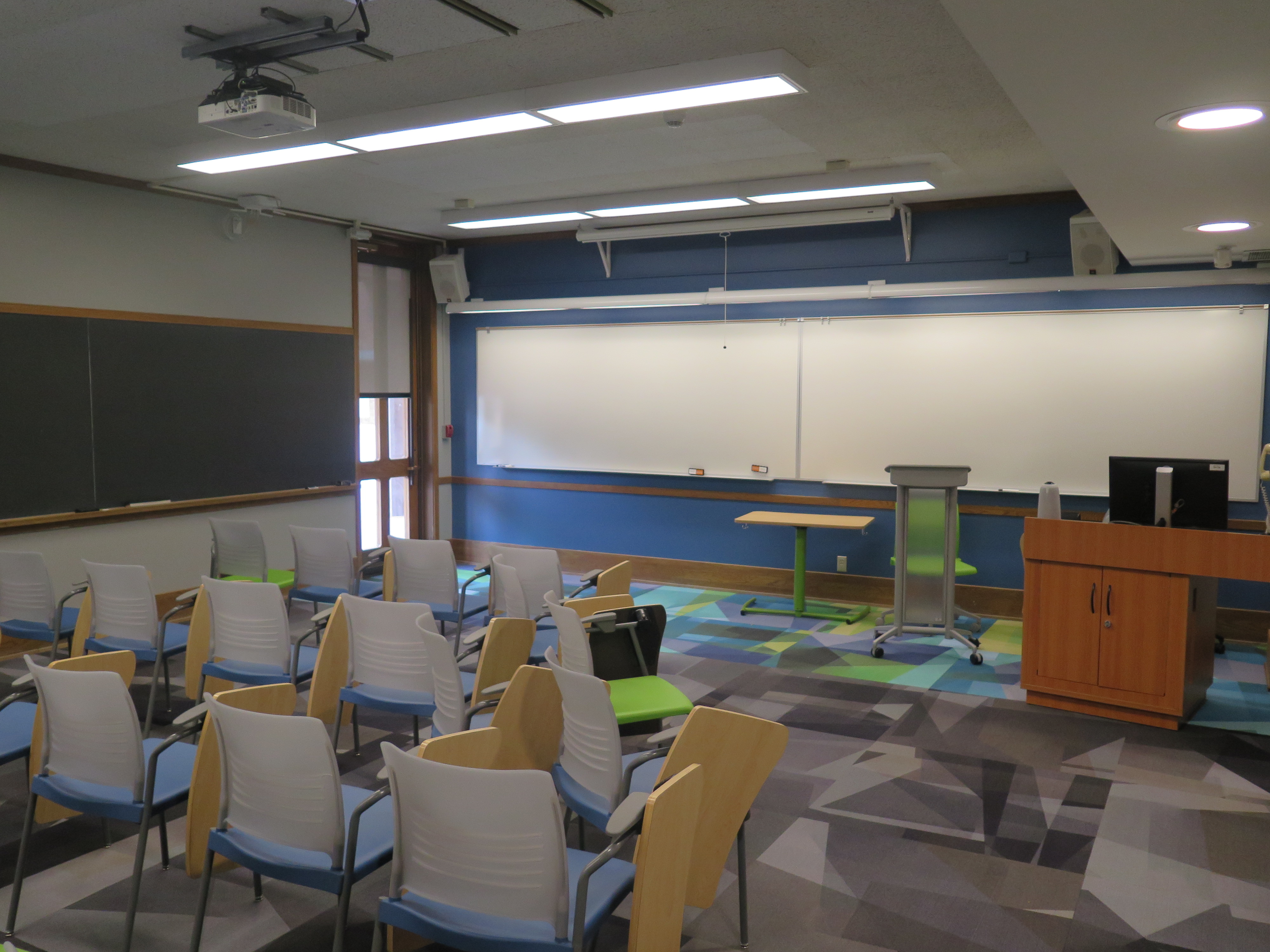 View from the classroom entrance. Consists of Moveable tablet arm chairs. Moveable Table and chair at the from of the room. Podium at the front of the room, white boards across the front wall of the room and chalkboards on the outer walls of the classroom. 