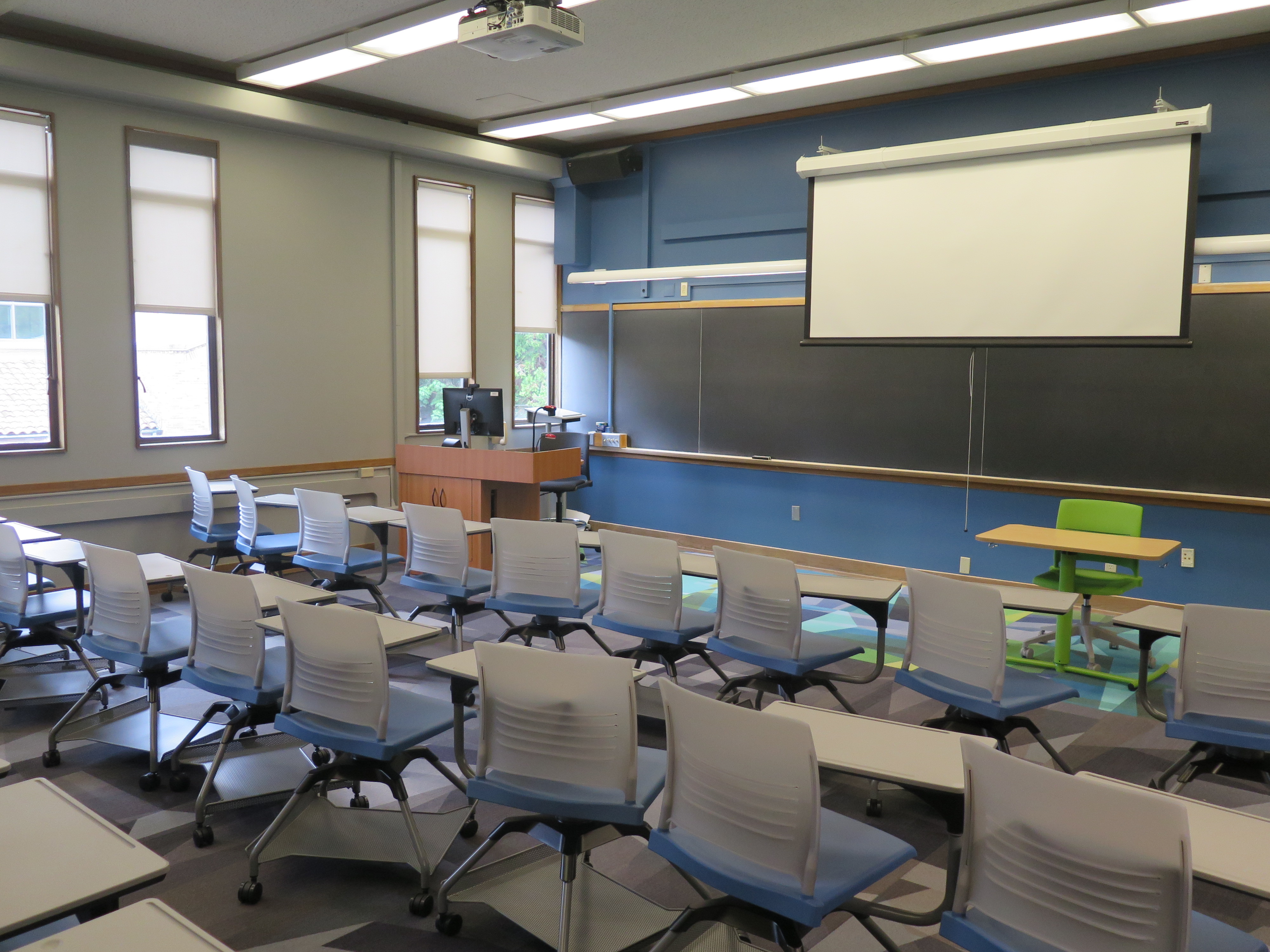View from the classroom entrance. Consists of Moveable tablet arm chairs. Moveable Table and chair at the from of the room. Podium and chalkboard at the front of the room 