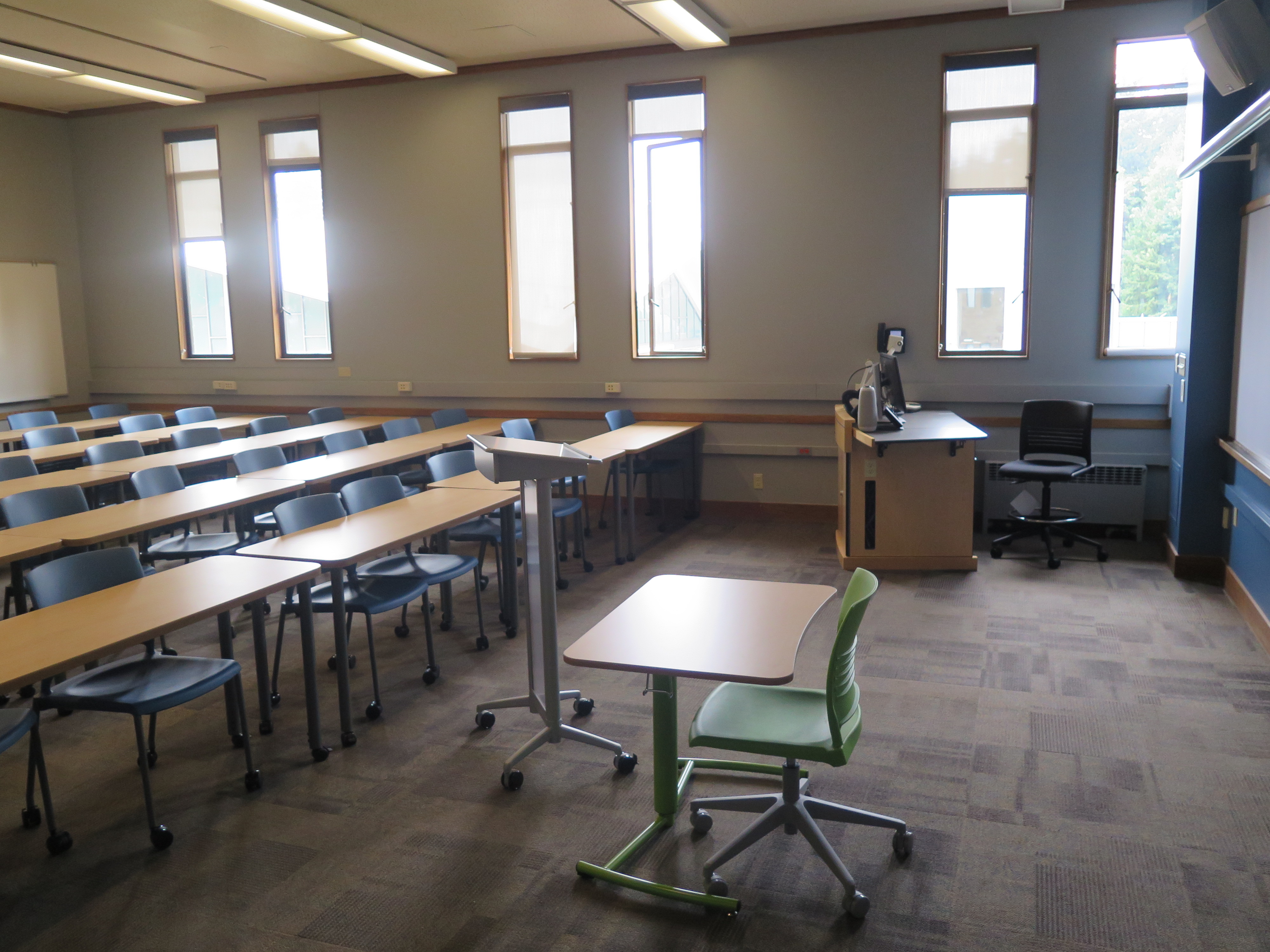 View from the classroom entrance. Consists of Moveable tables and chairs. Podium at the front of the room, white boards across the front and back walls.