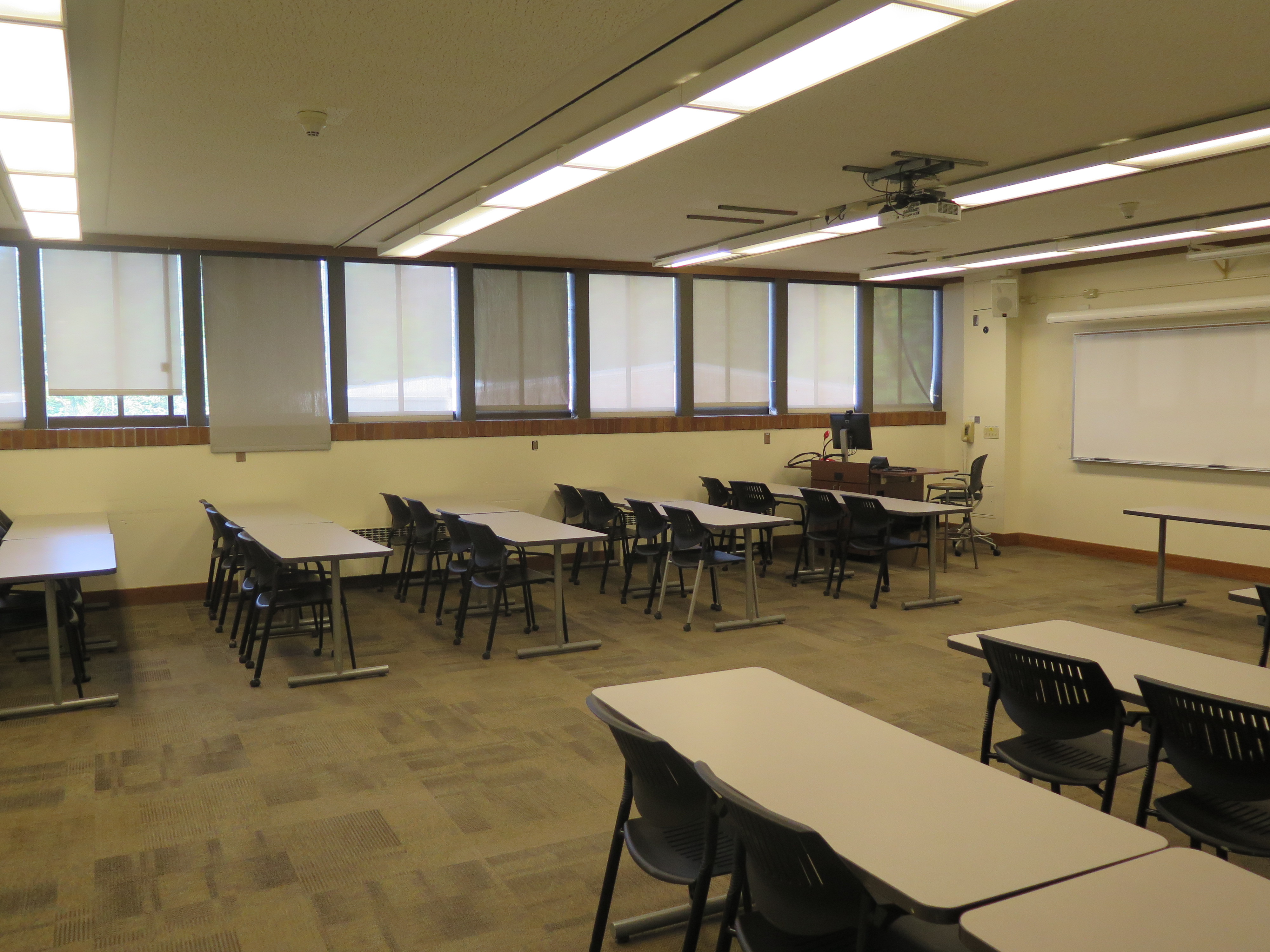 This room contains carpet floor, moveable tables and chairs, a whiteboard at the front of the room and a podium.