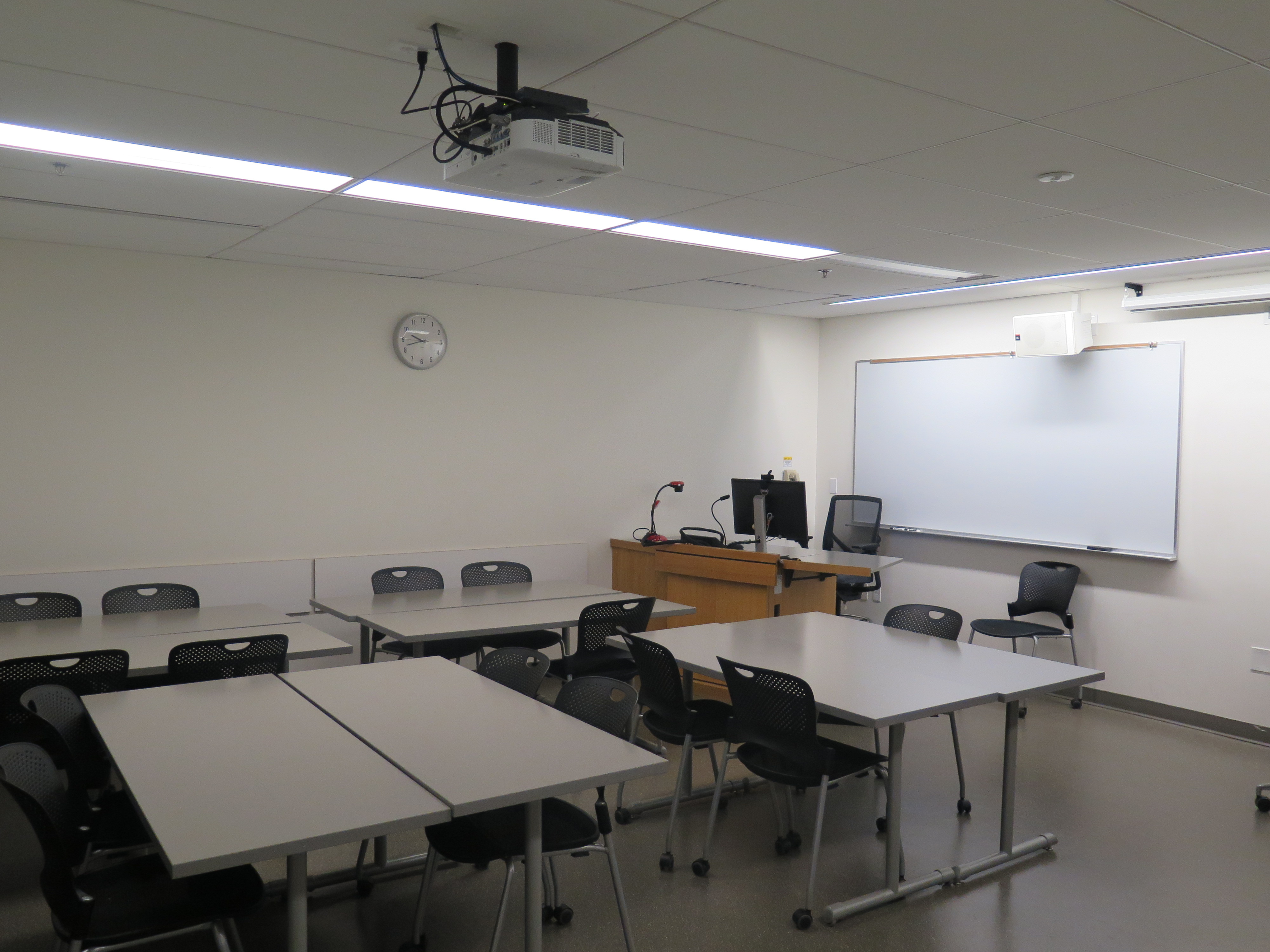 Room Consists of hard floors, moveable tables and chairs, a white board and podium are both located at the front of the room.
