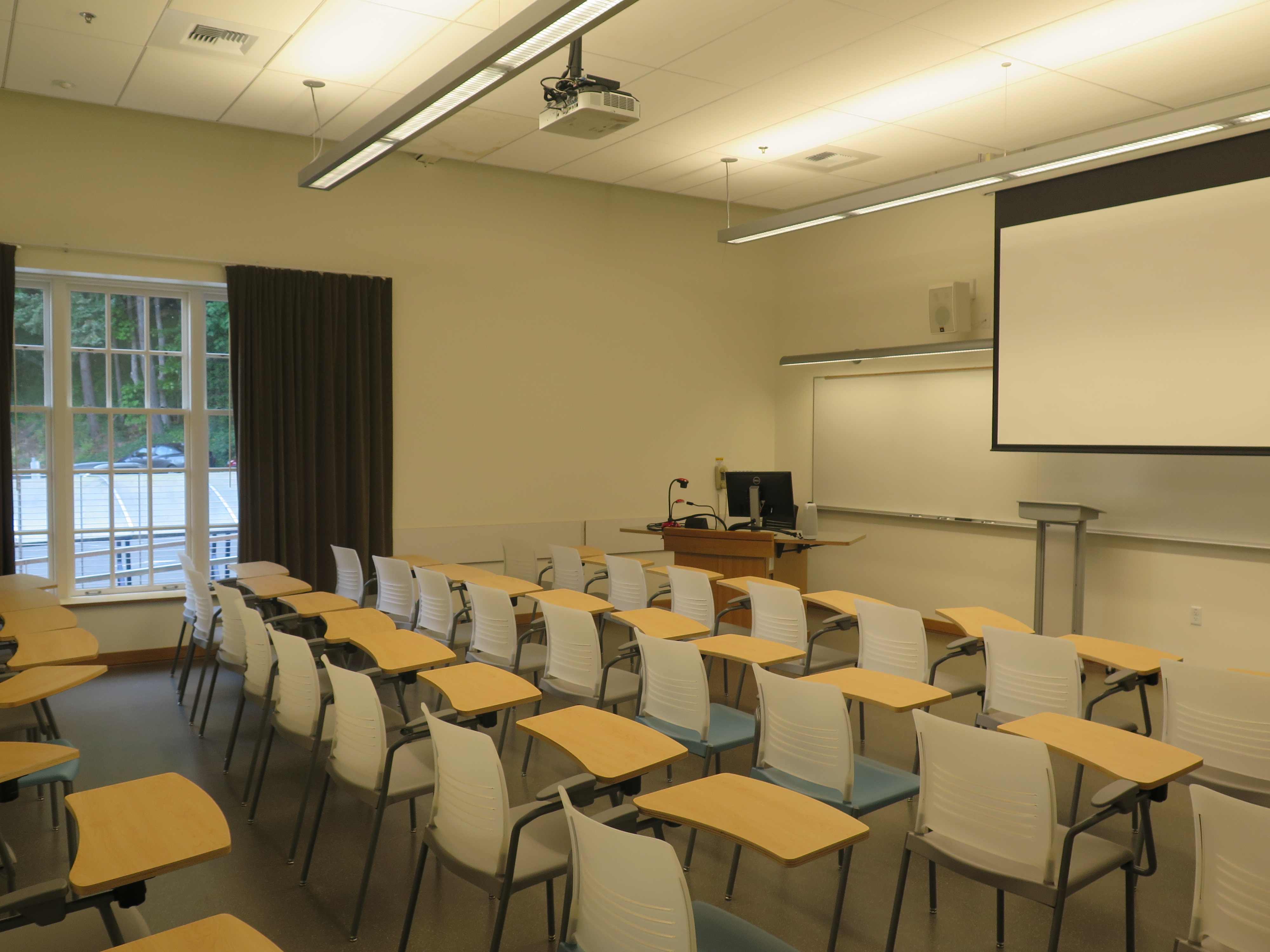 Room Consists of hard floors, moveable tablet armchairs, a white board and podium are both located at the front of the room with another whiteboard located on the right wall of the room.