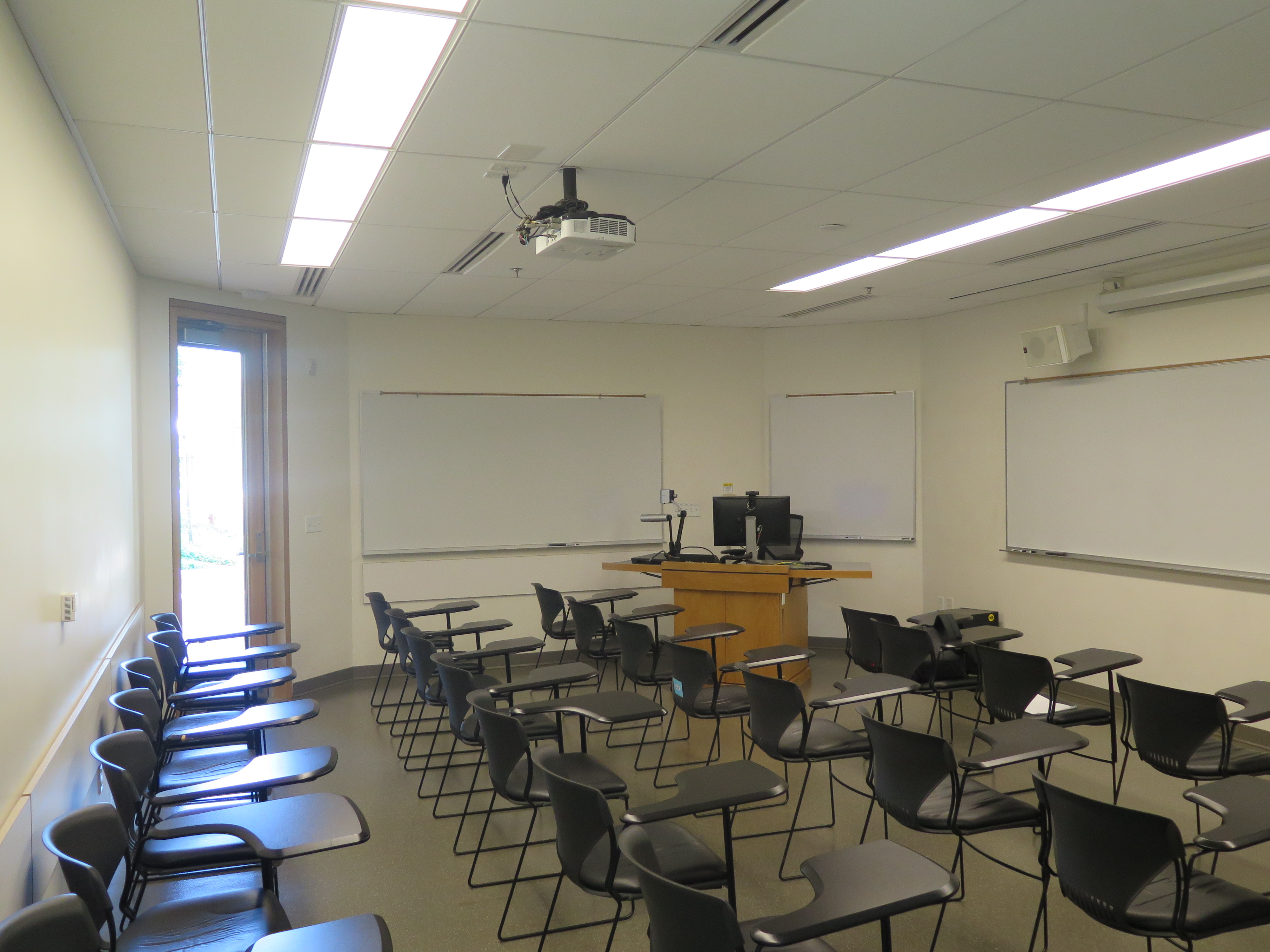 Room Consists of hard floors, moveable tablet armchairs, a white board and podium are both located at the front of the room and whiteboards on the outer walls of the room.