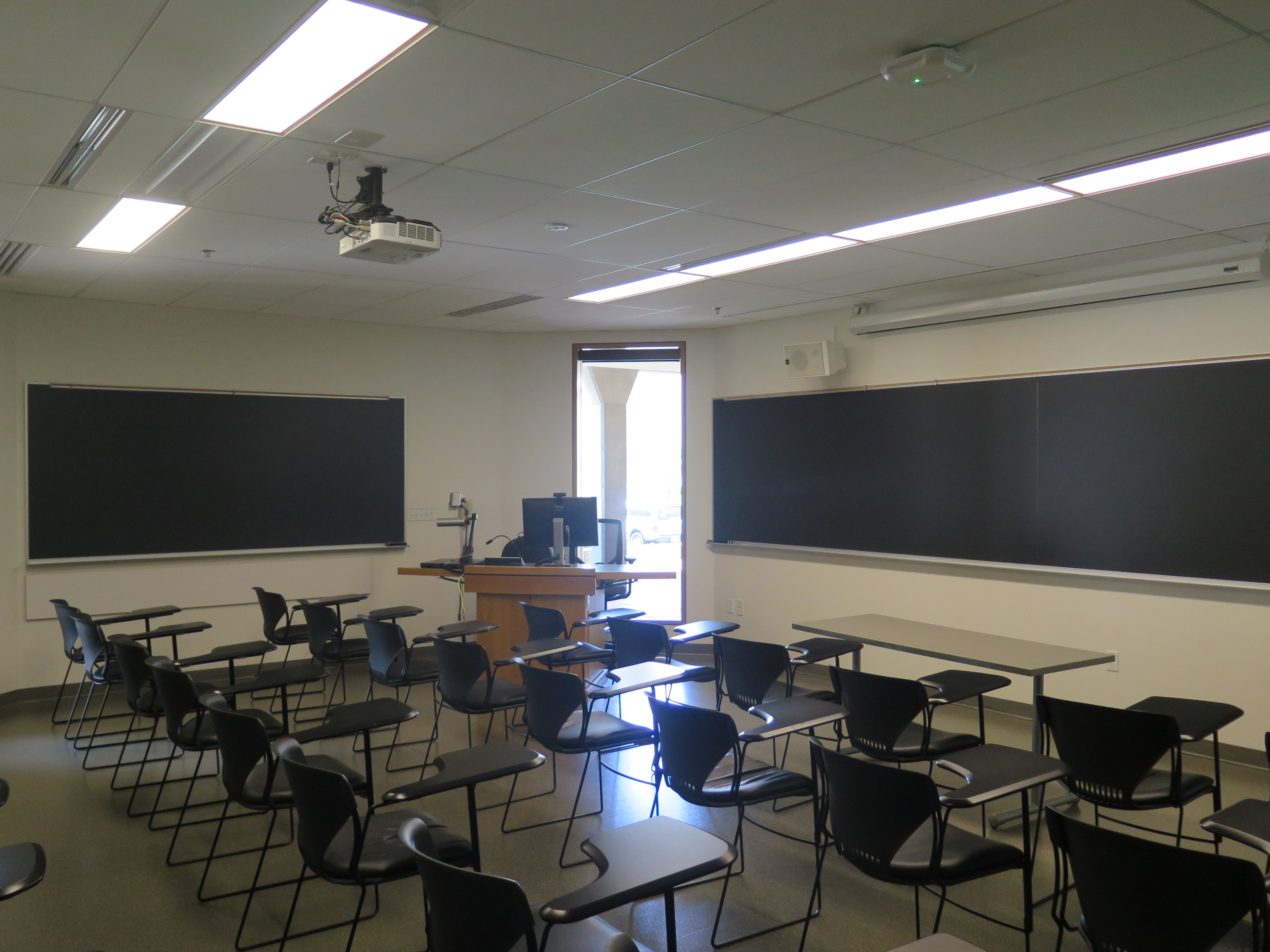 Room Consists of hard floors, moveable tablet armchairs, a chalk board and podium are both located at the front of the room with chalk boards located on the outer walls.