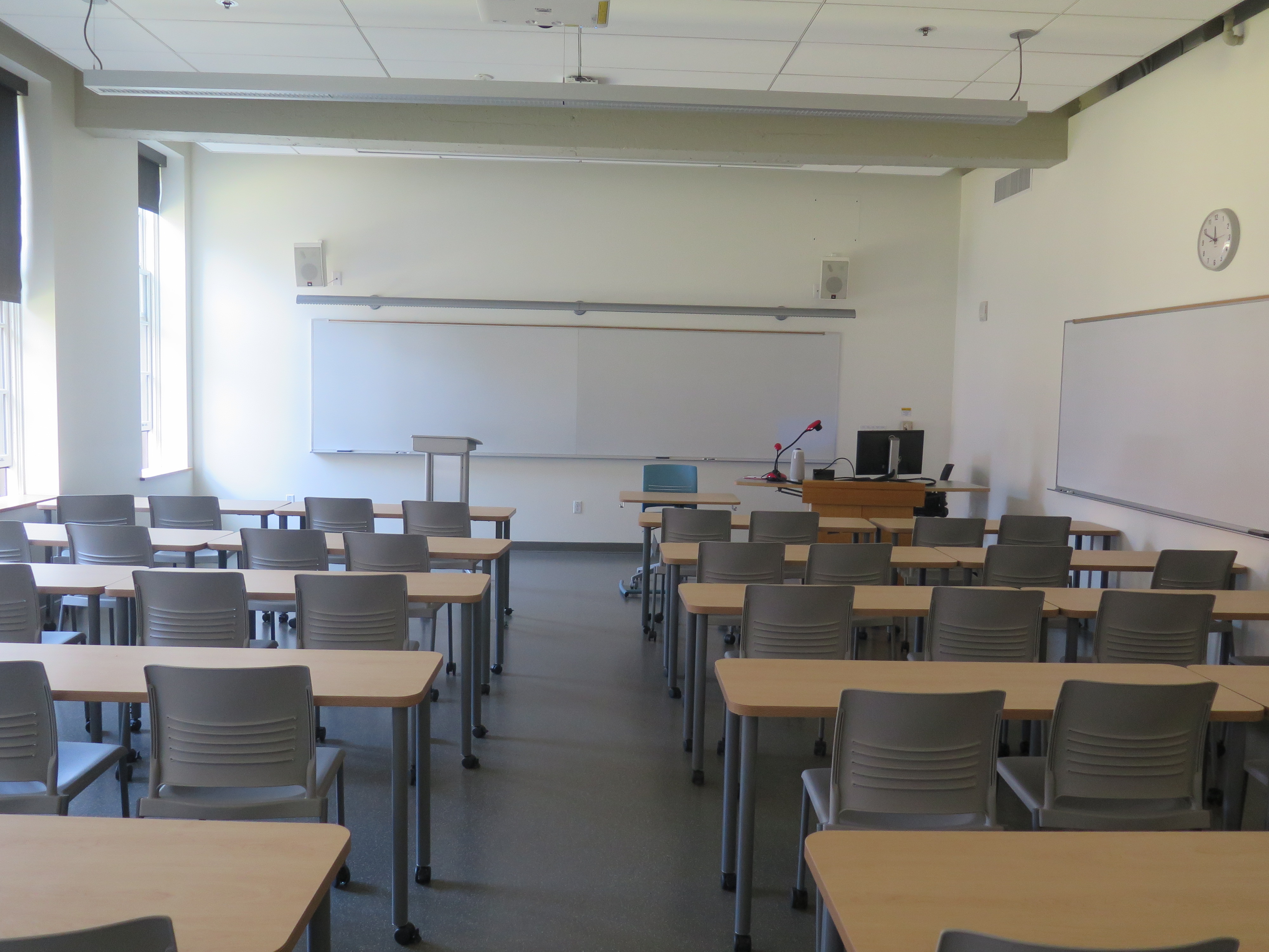 Room Consists of hard floors, moveable tables and chairs, a white board and podium are both located at the front of the room with another white board located on the right outer wall of the room.