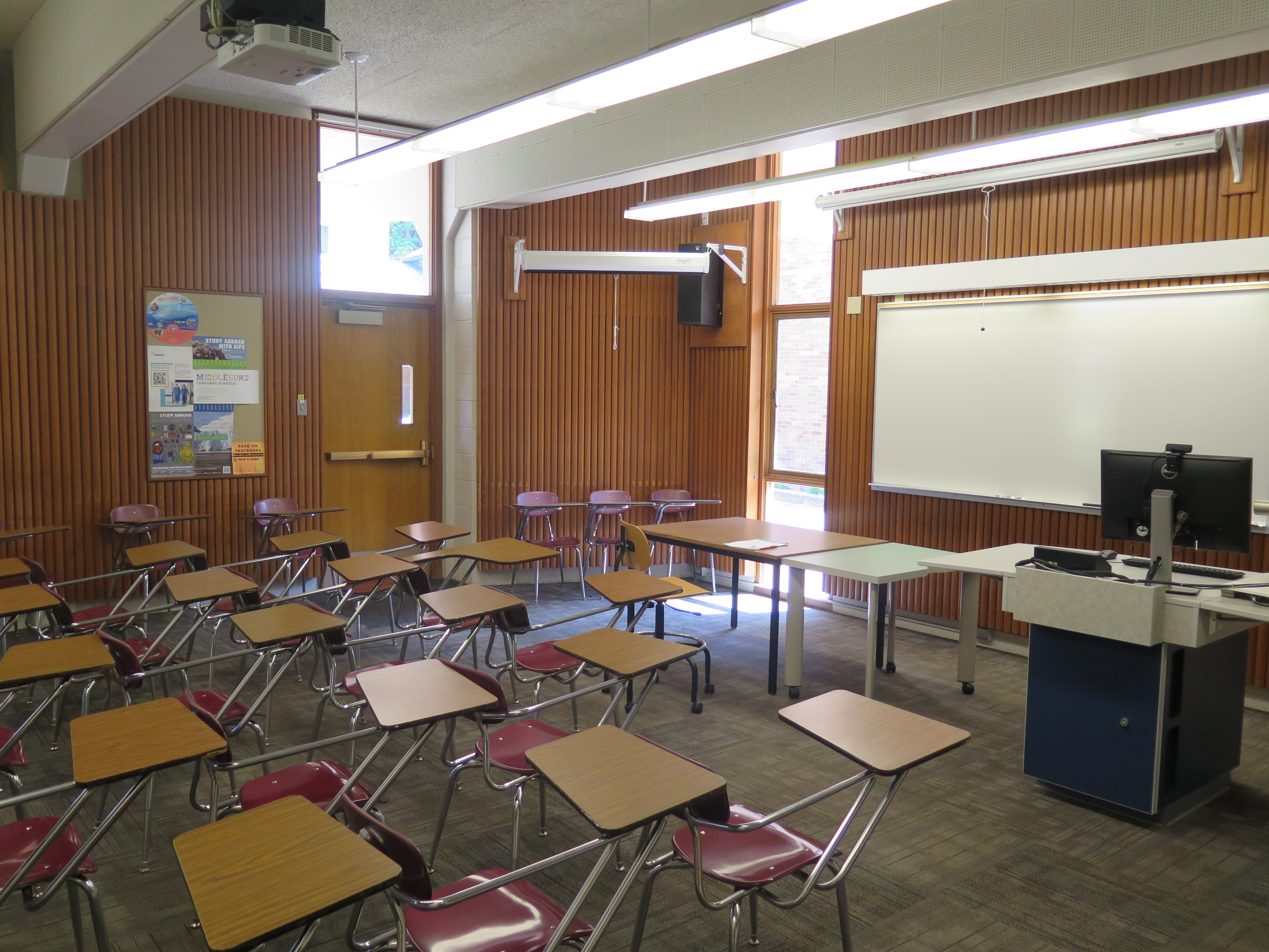 This room contains carpet floor, woods desks connected to chairs, a whiteboard at the front of the room and a podium 