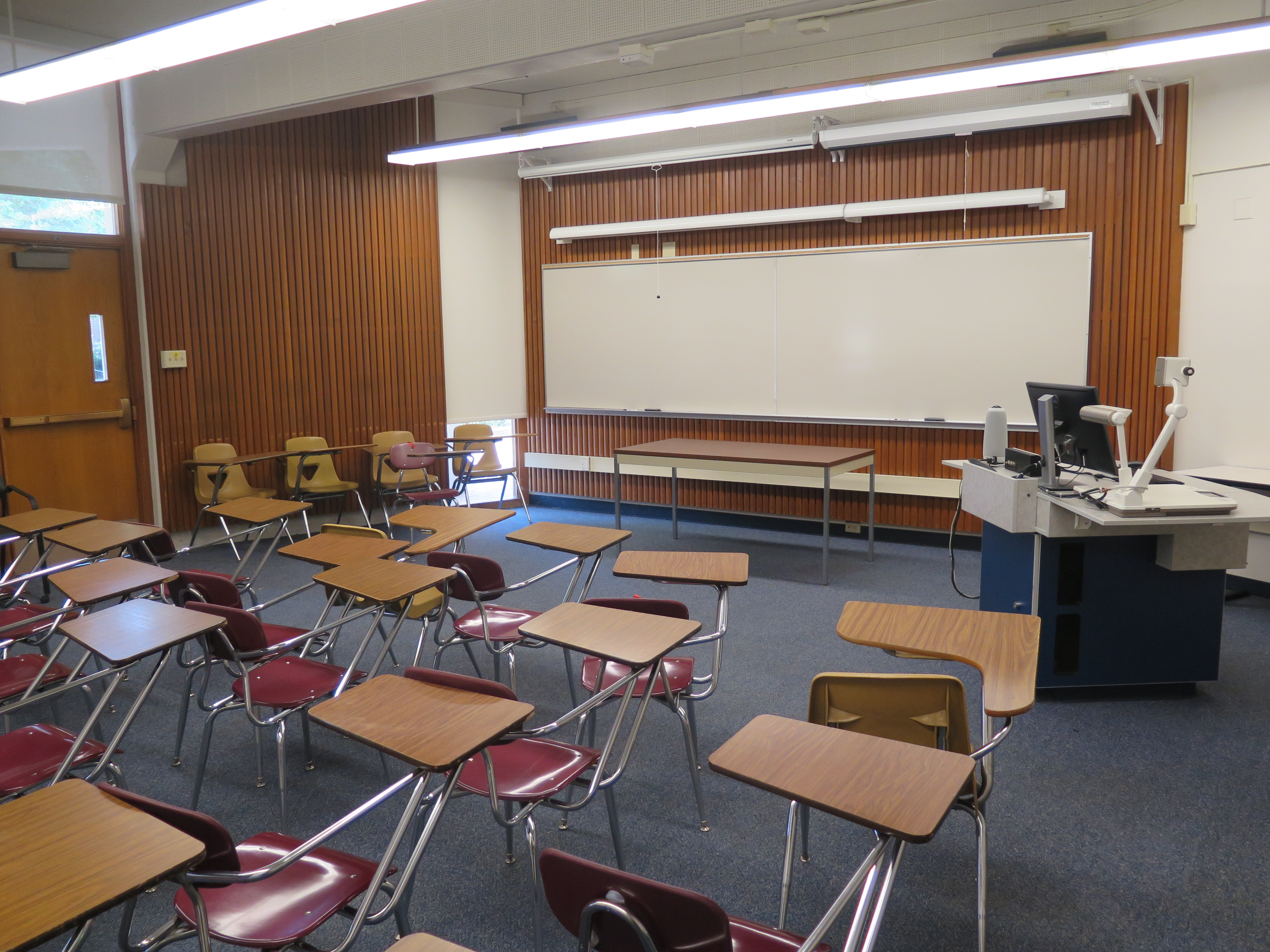 This room contains carpet floor, woods desks connected to chairs, a whiteboard at the front of the room and a podium 