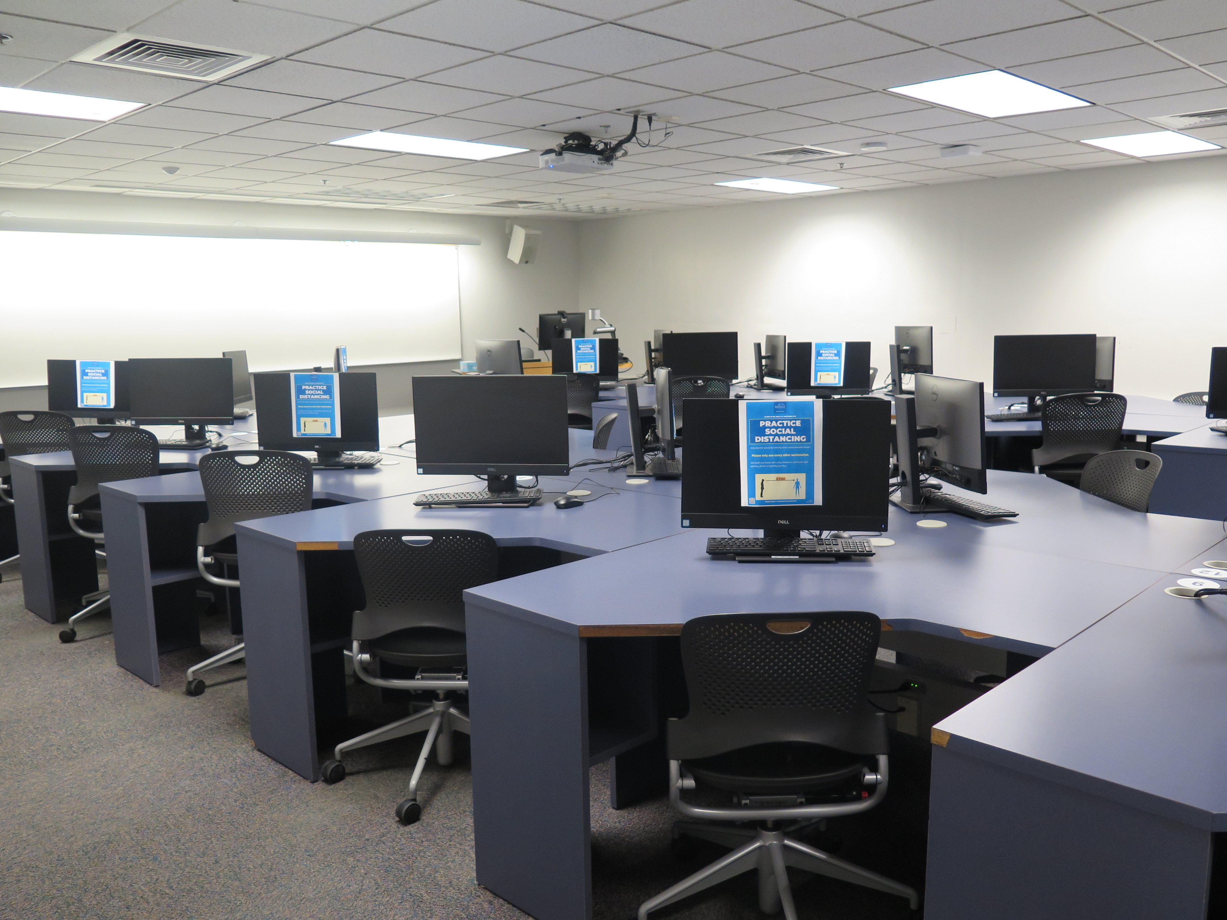 Computer Lab consists of carpet floor, individual computer stations in a row with an aisle in between each row, and Podium and whiteboard located at the front of the room.
