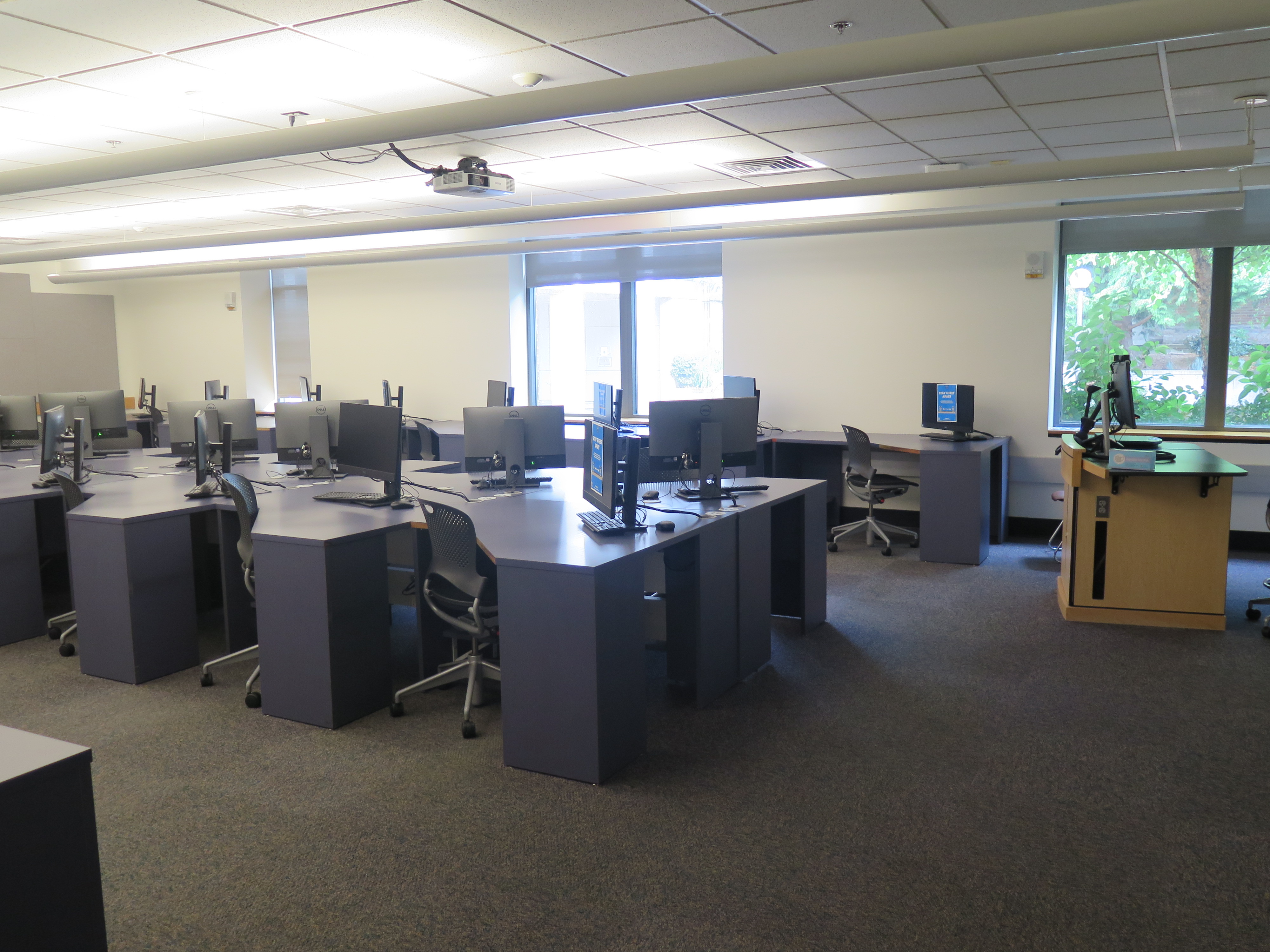 Computer Lab consists of carpet floor, individual computer stations in a row with an aisle in between each row, and Podium and whiteboard located at the front of the room.
