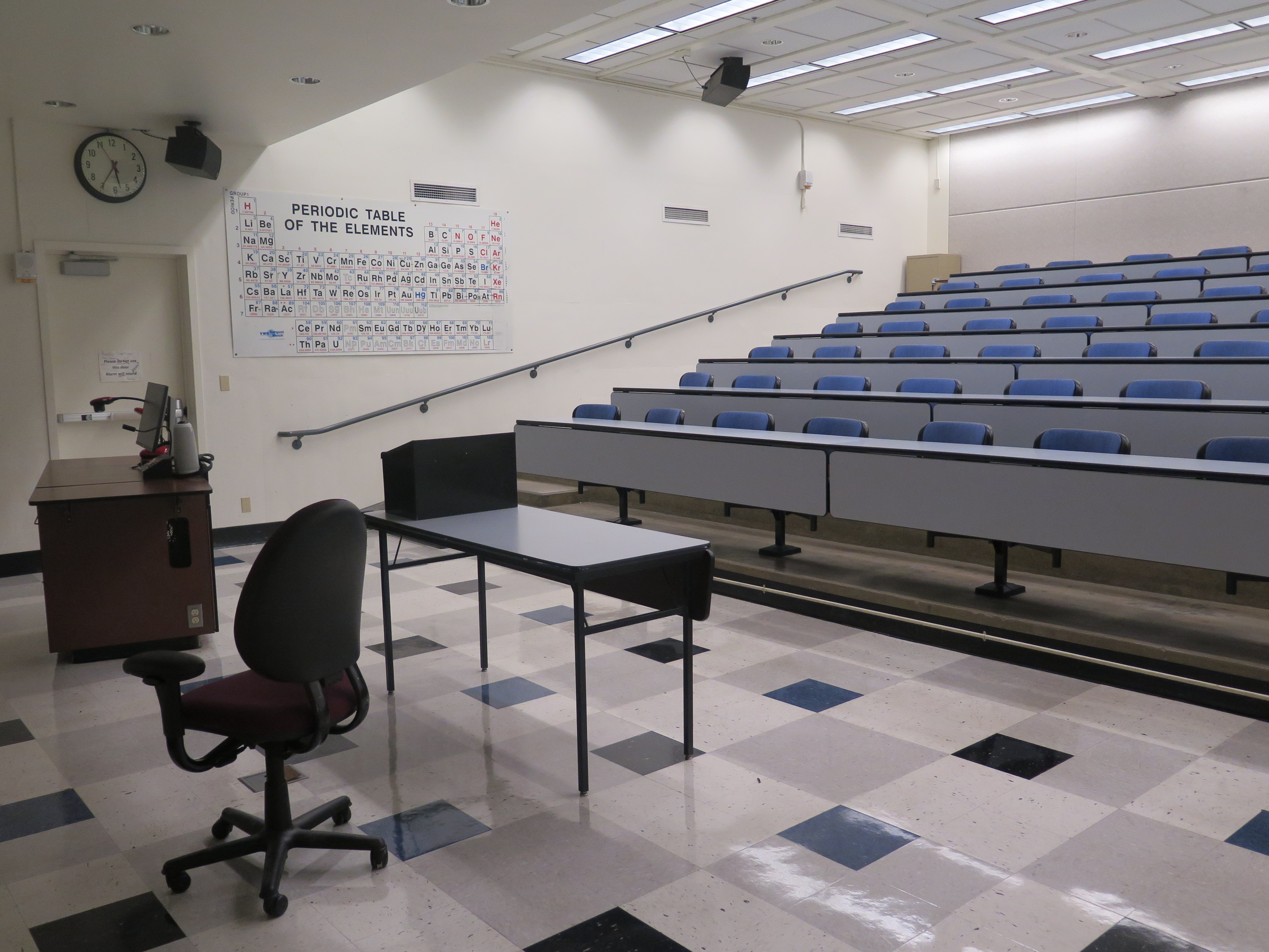 Room Consists of hard floors, Stationary rows of tables and chairs, a white board and podium are both located at the front of the room. This room also uses a Rear Screen projector, it is the large Gray surface next to the white board