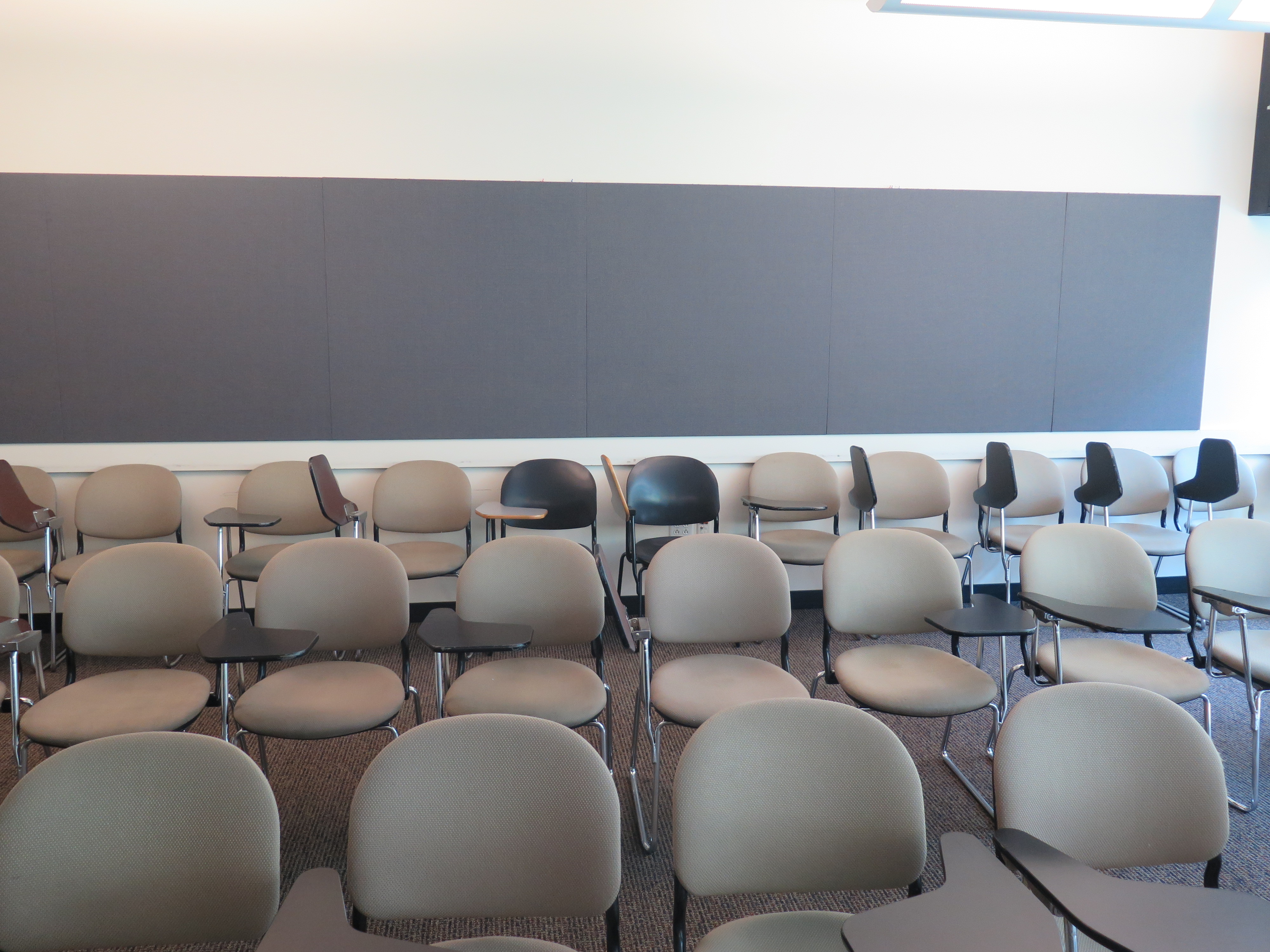 Room consists of carpet, moveable tablet armchairs and a table, podium and whiteboard located at the front of the room. 