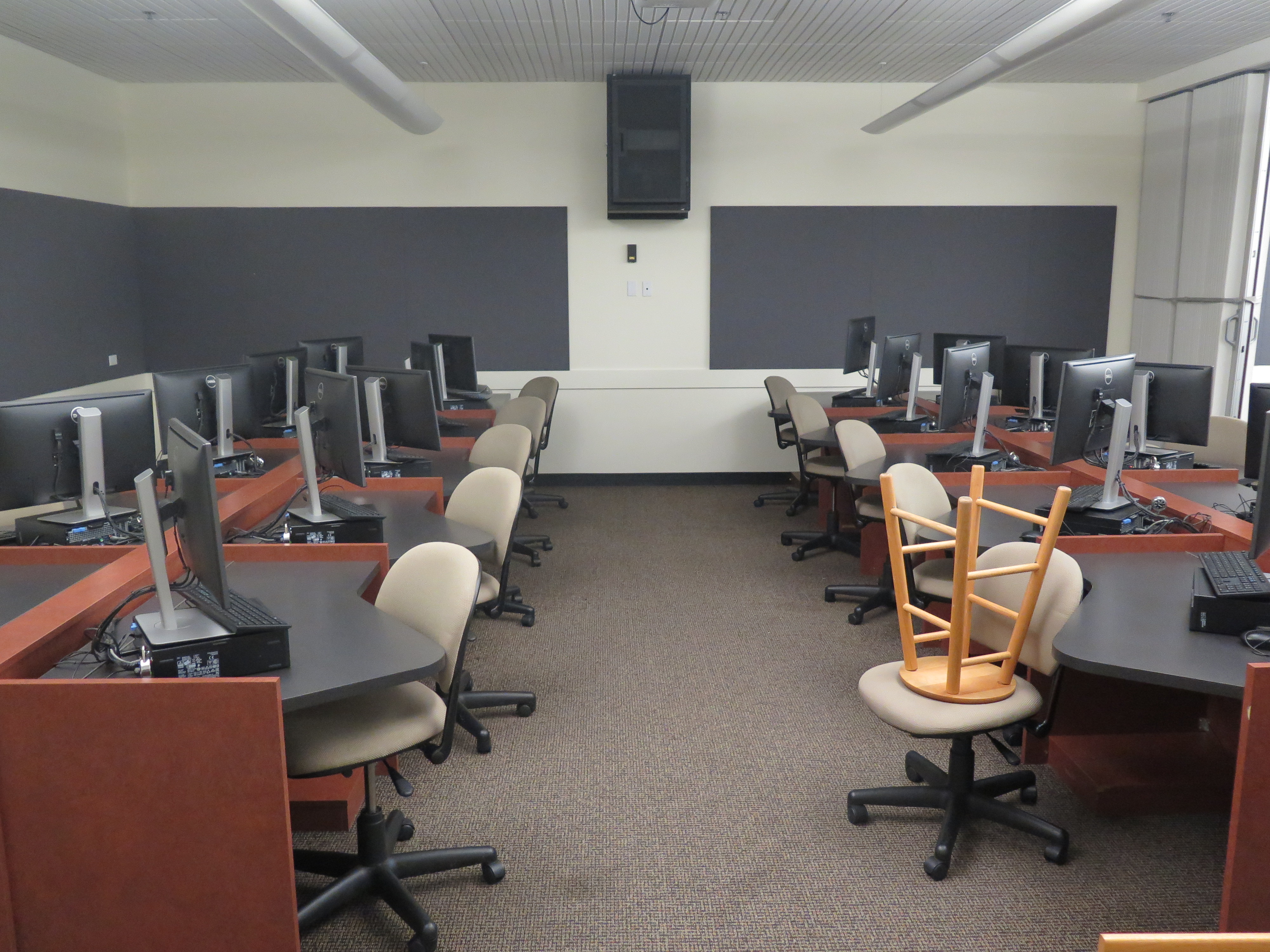 Computer Lab consists of carpet floor, individual computer stations in a row with an aisle in between each row, and Podium and whiteboard located at the front of the room. 