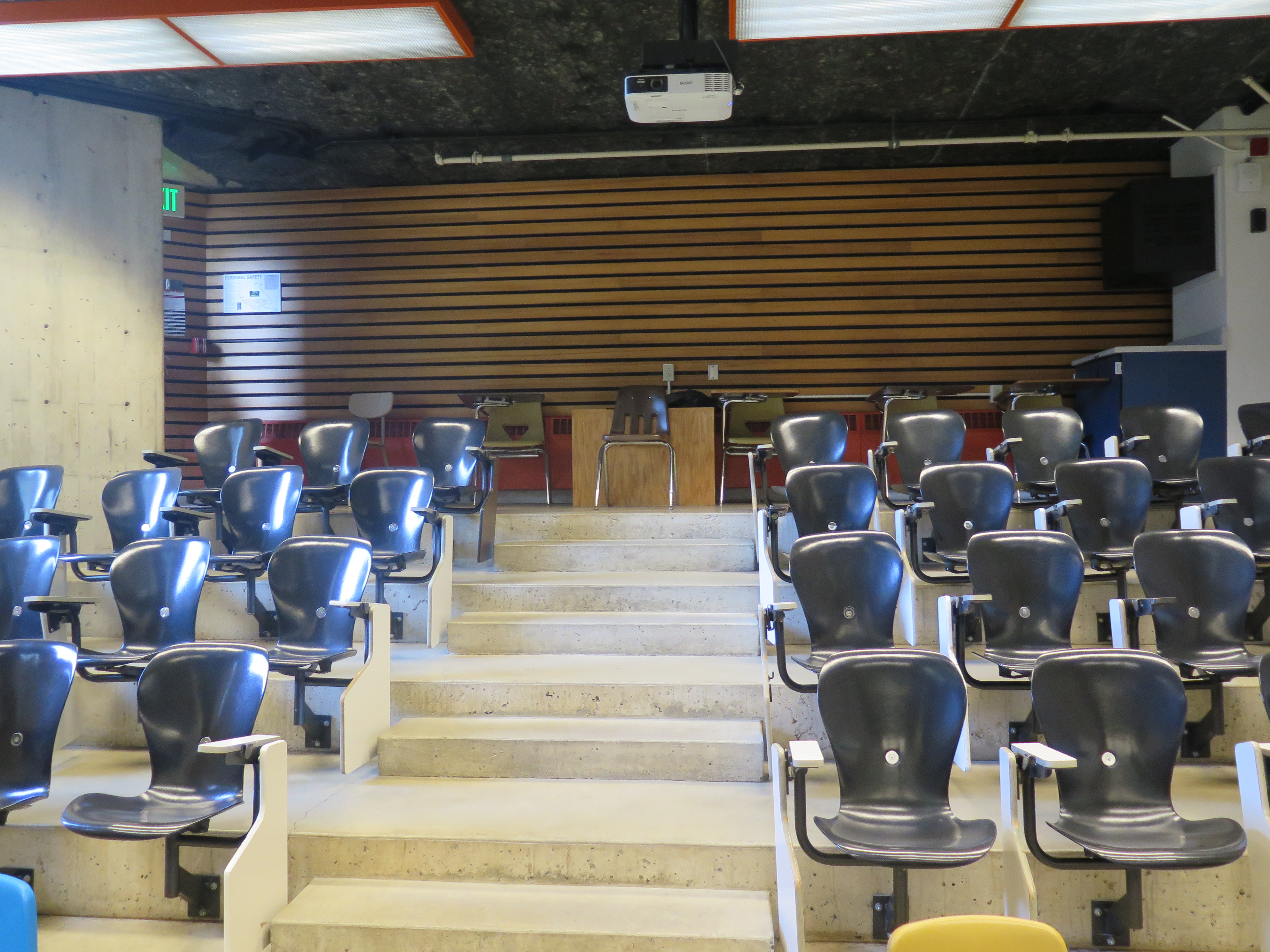 Room Consists of auditorium style, stationary tablet armchairs, White board and podium are located at the front of the room.