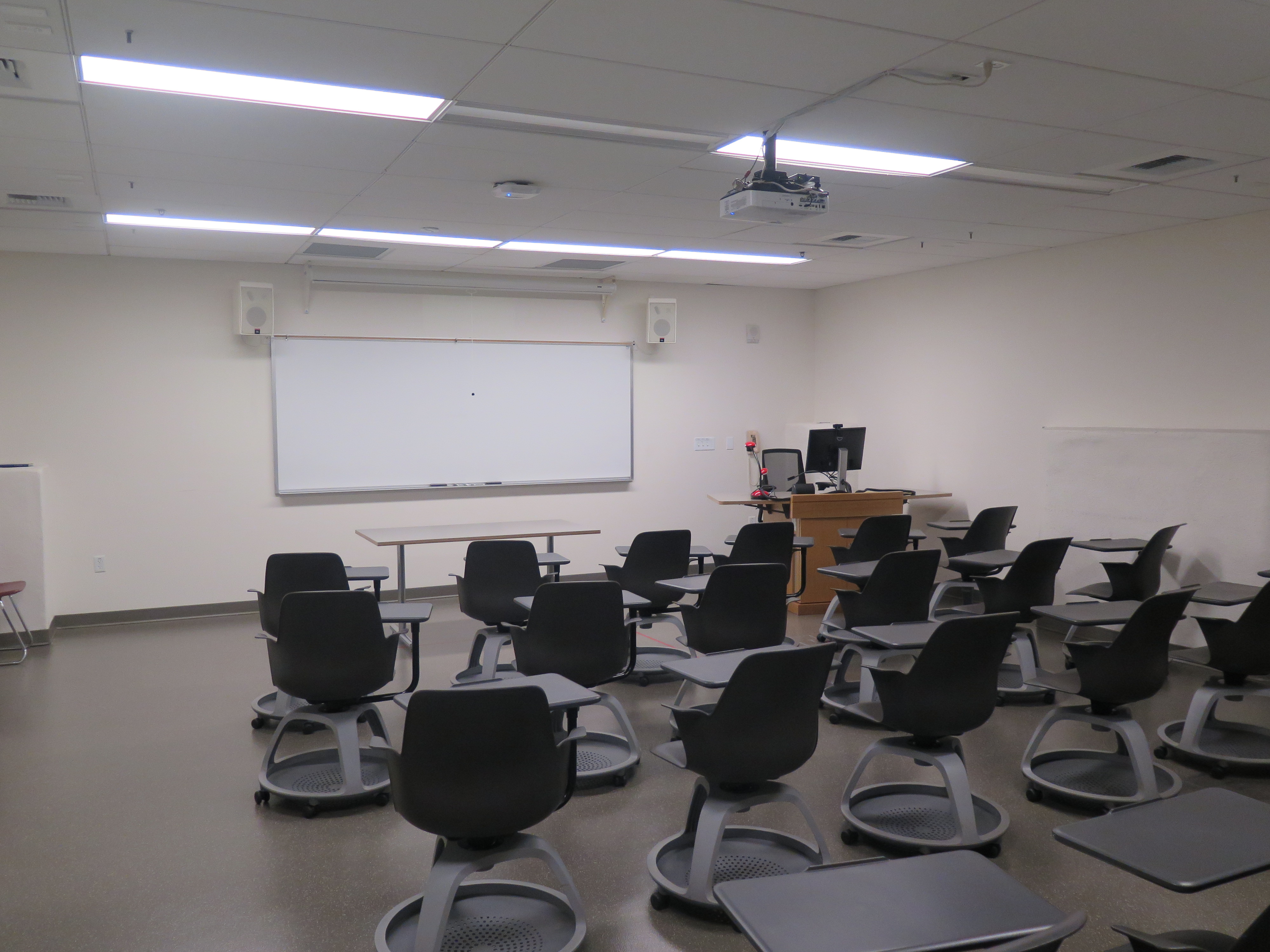 Room Consists of hard floors, moveable tablet armchairs, a white board and podium are both located at the front of the room.