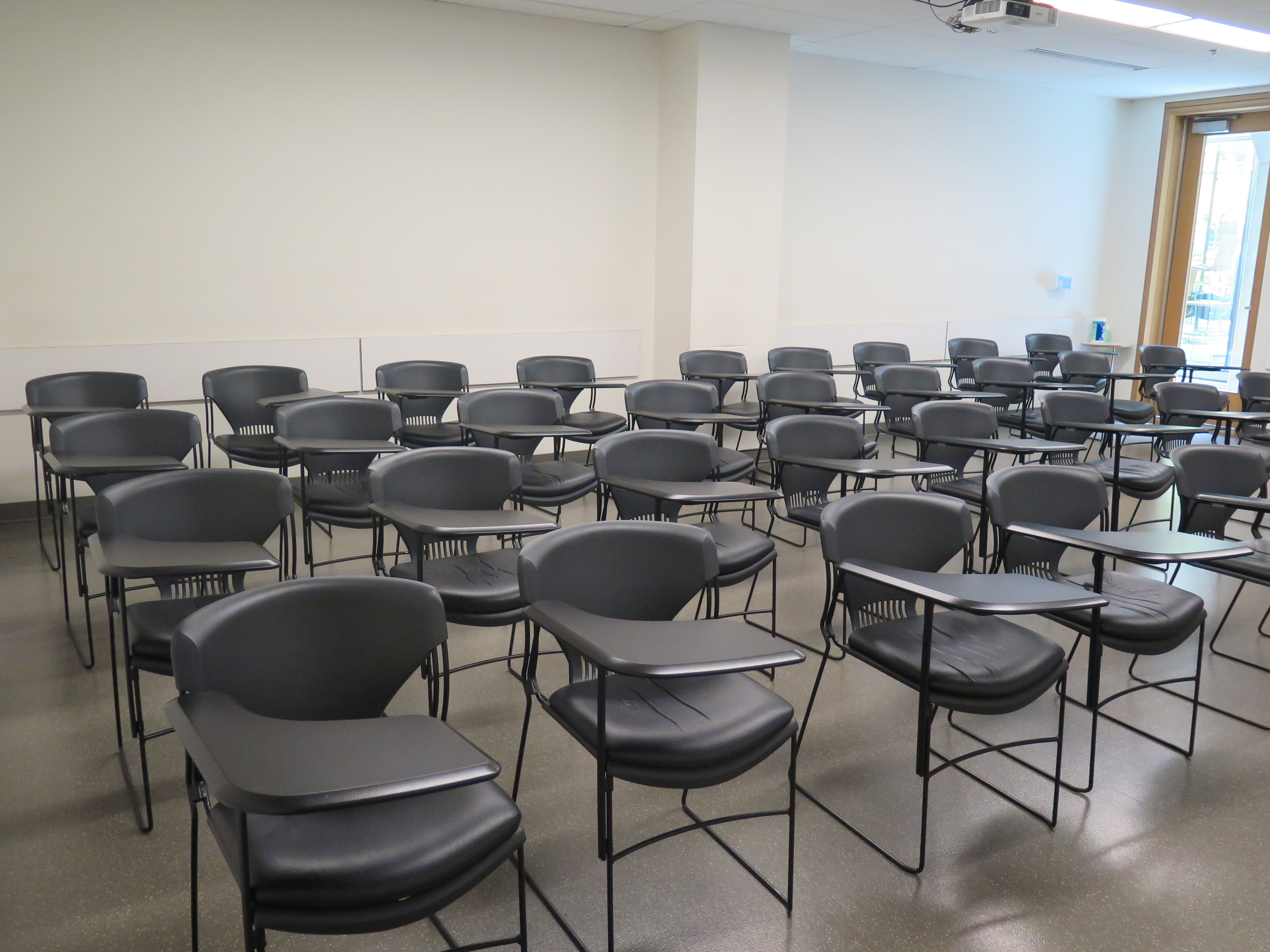 Room Consists of hard floors, moveable tablet armchairs, a white board and podium are both located at the front of the room.