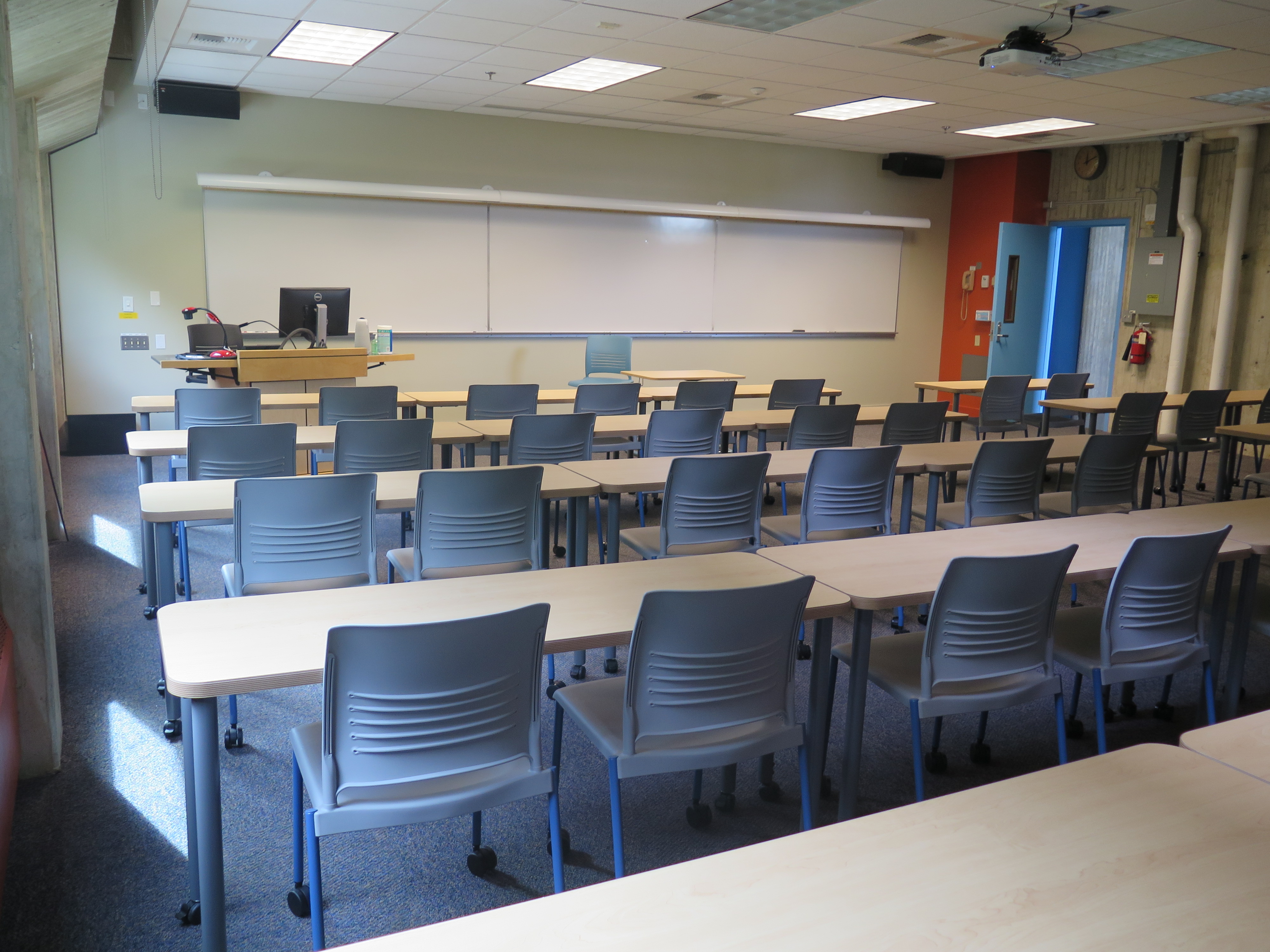 Room consists of carpet, moveable tables and chairs and a podium and whiteboard located at the front of the room. 