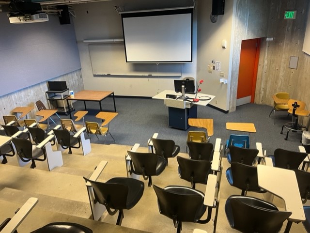 Overall view of the room, it has fixed tablet arm chairs, whiteboard Tiered seating and windows on the south side of the room