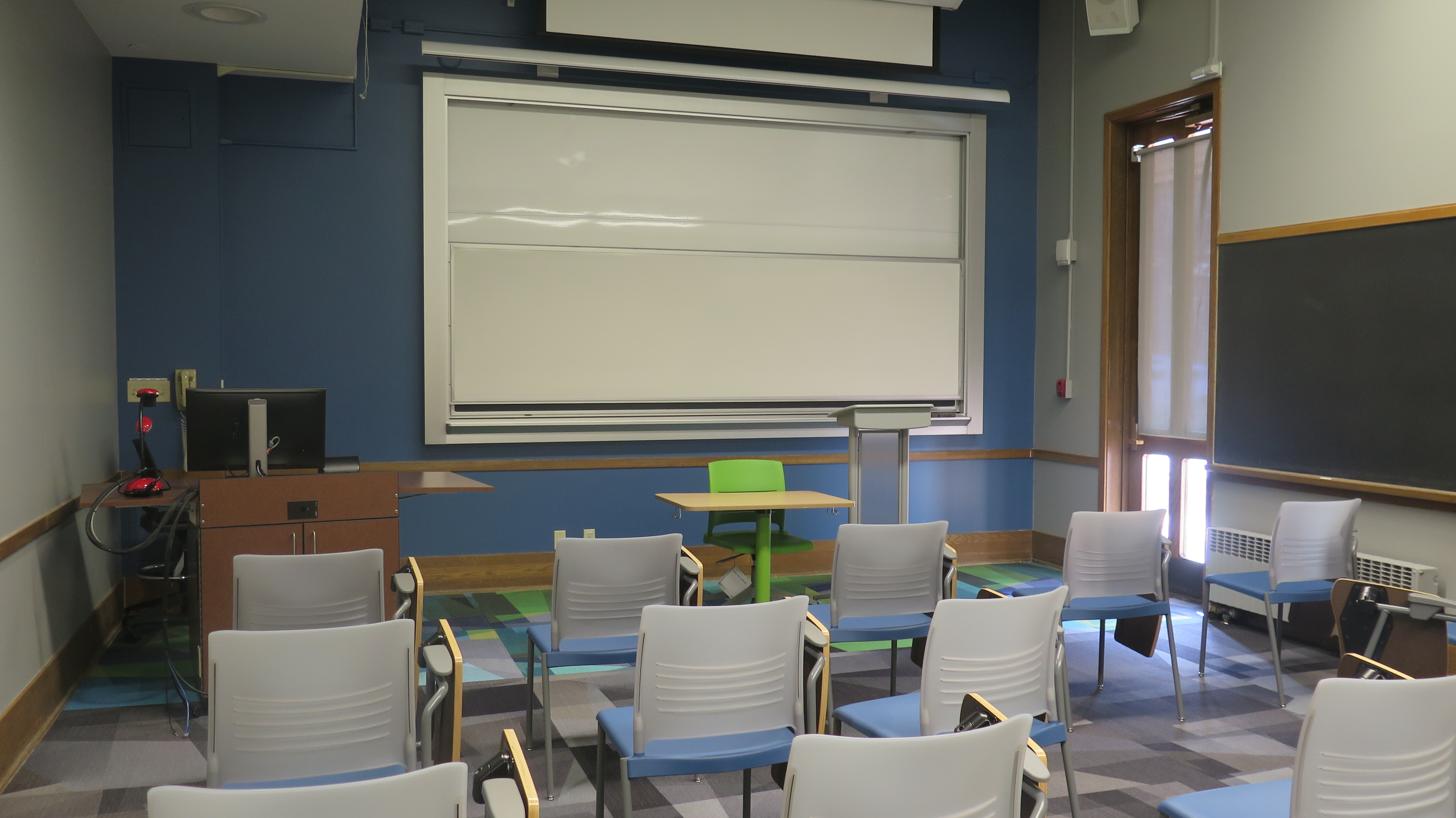 View from the back of the room, Carpet, Moveable tablet arm chairs and podium. Whiteboard on the front wall of classroom
