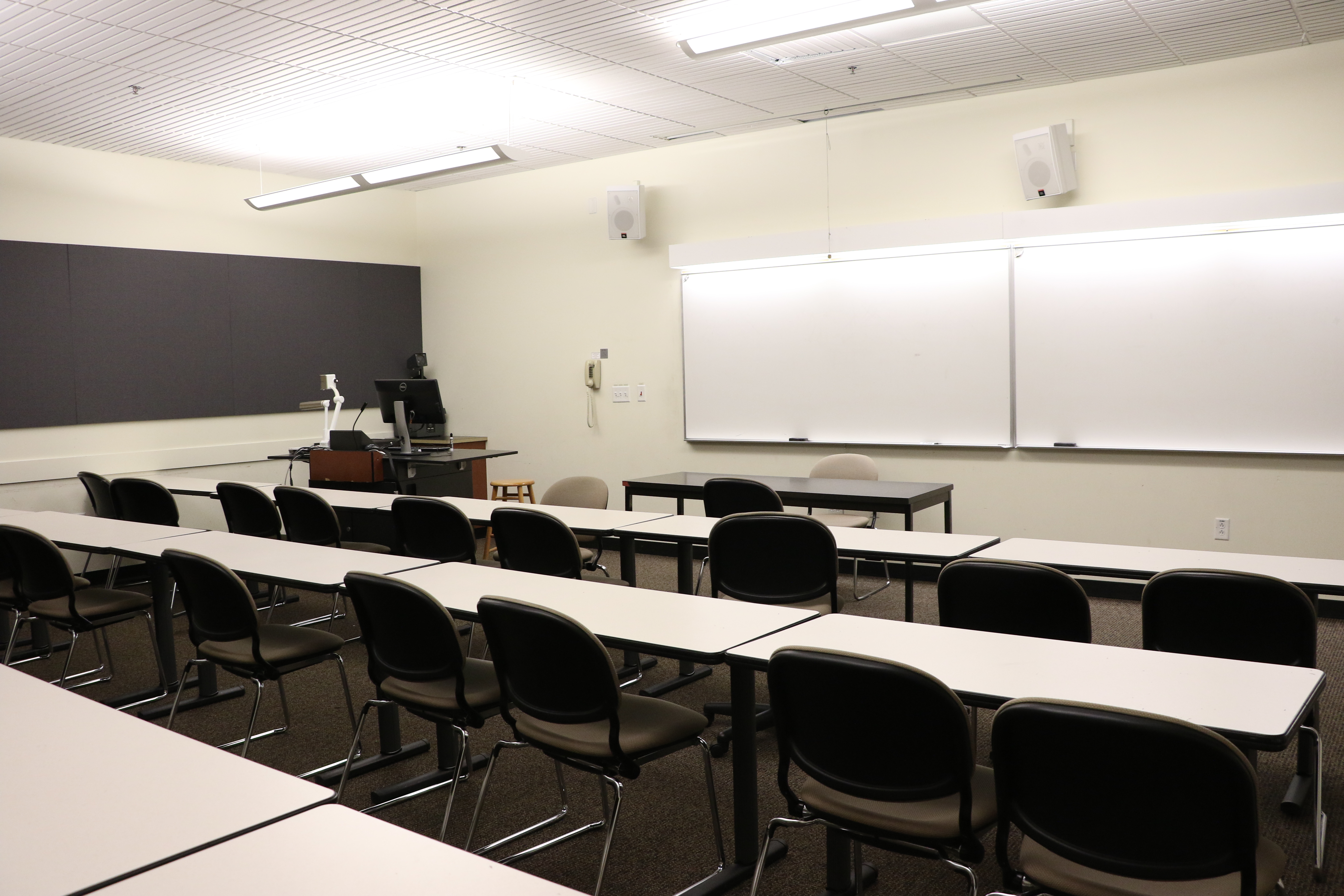 Room consists of carpet, moveable tables and chairs and a table, podium and whiteboard located at the front of the room. 