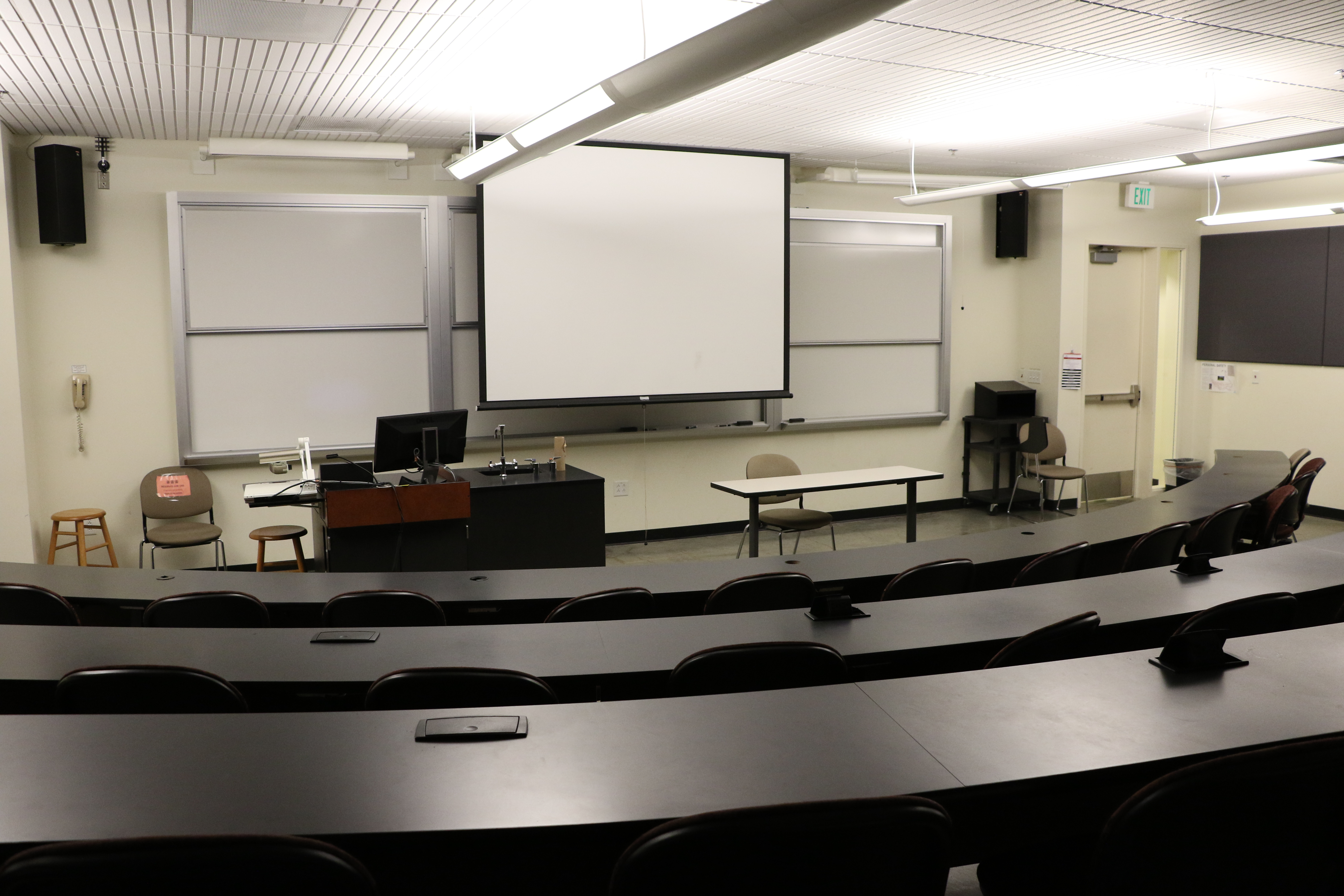 Room Consists of hard floors, Stationary rows of tables and chairs, a white board and podium are both located at the front of the room.