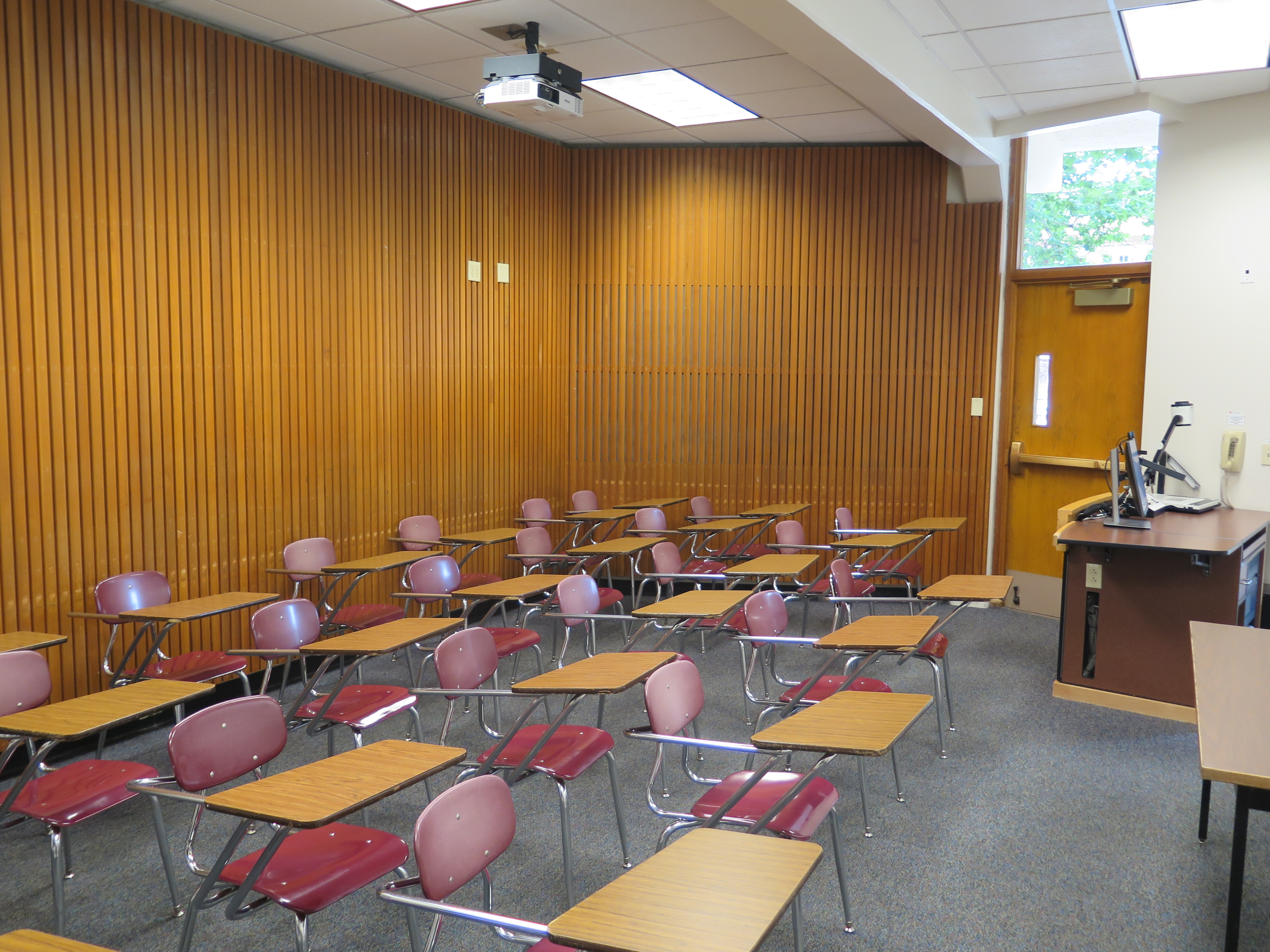 Room Consists of Carpet floors, moveable tables and chairs, a white board and podium are both located at the front of the room.