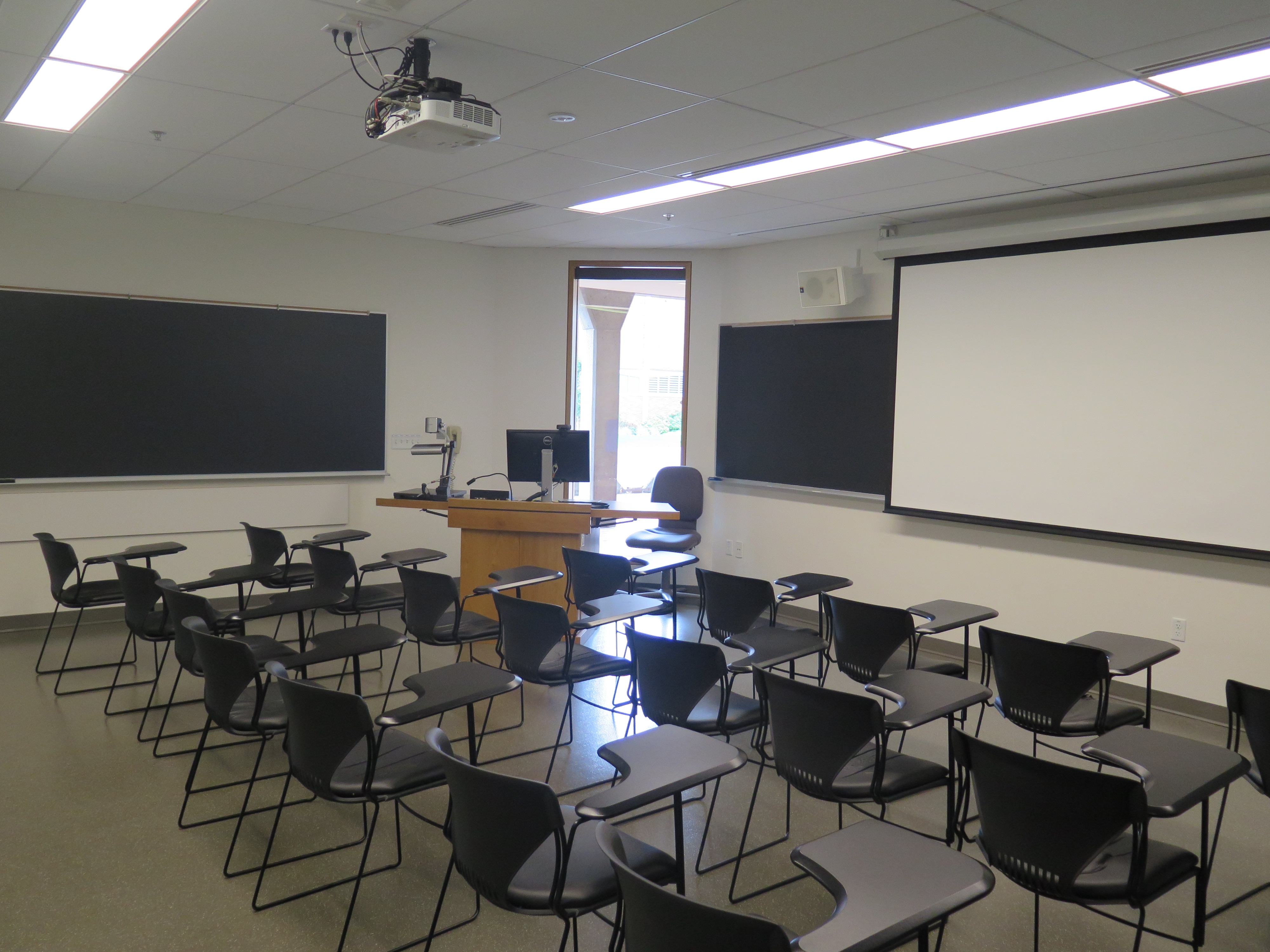 Room Consists of hard floors, moveable tablet armchairs, a white board and podium are both located at the front of the room and chalkboards are located on the outer right and left walls of the room.