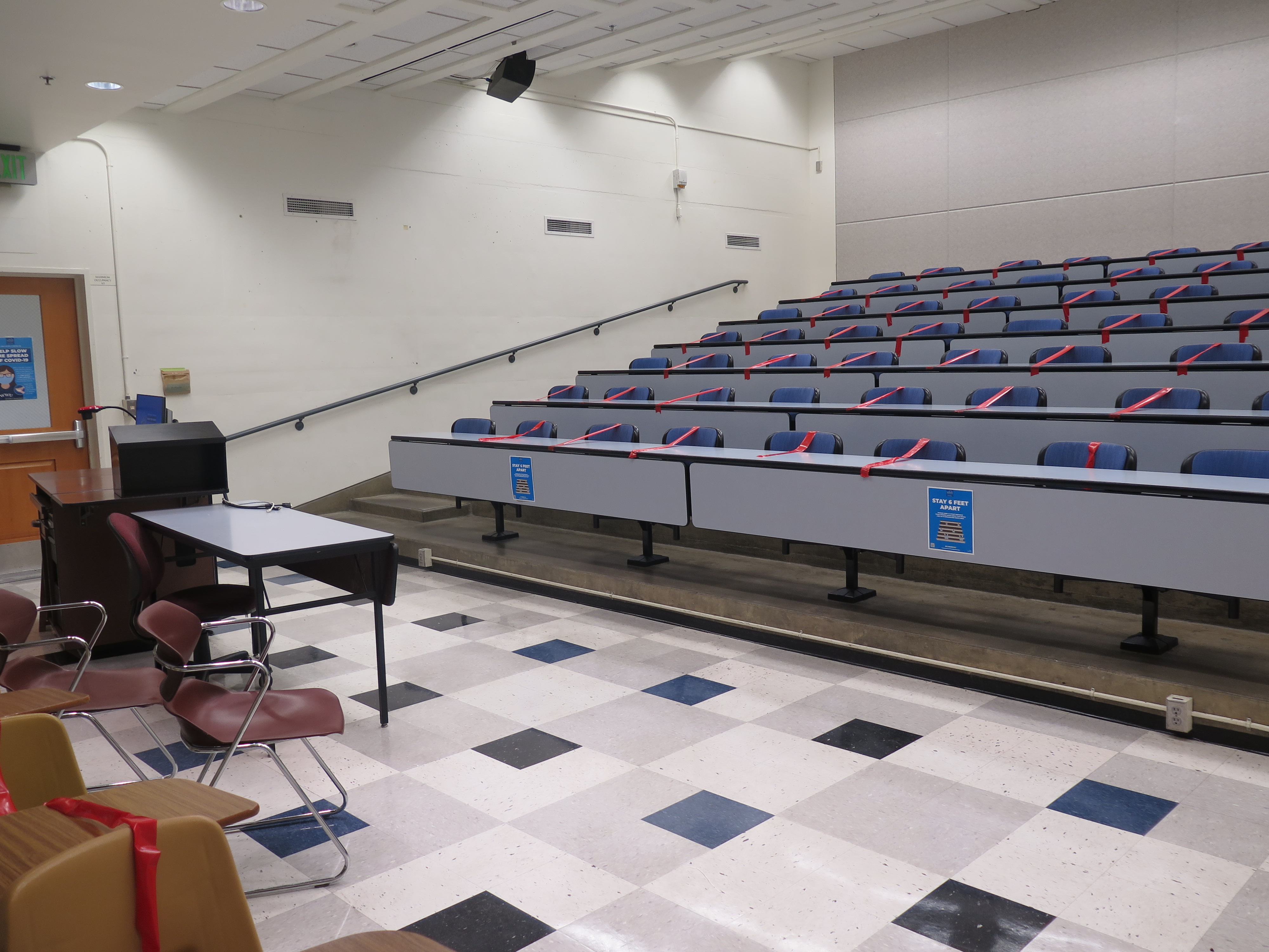 Room Consists of hard floors, Stationary rows of tables and chairs, a podium and whiteboard are located at the front of the room. This room uses a Rear Screen projector, it is the large Gray surface next to the white board