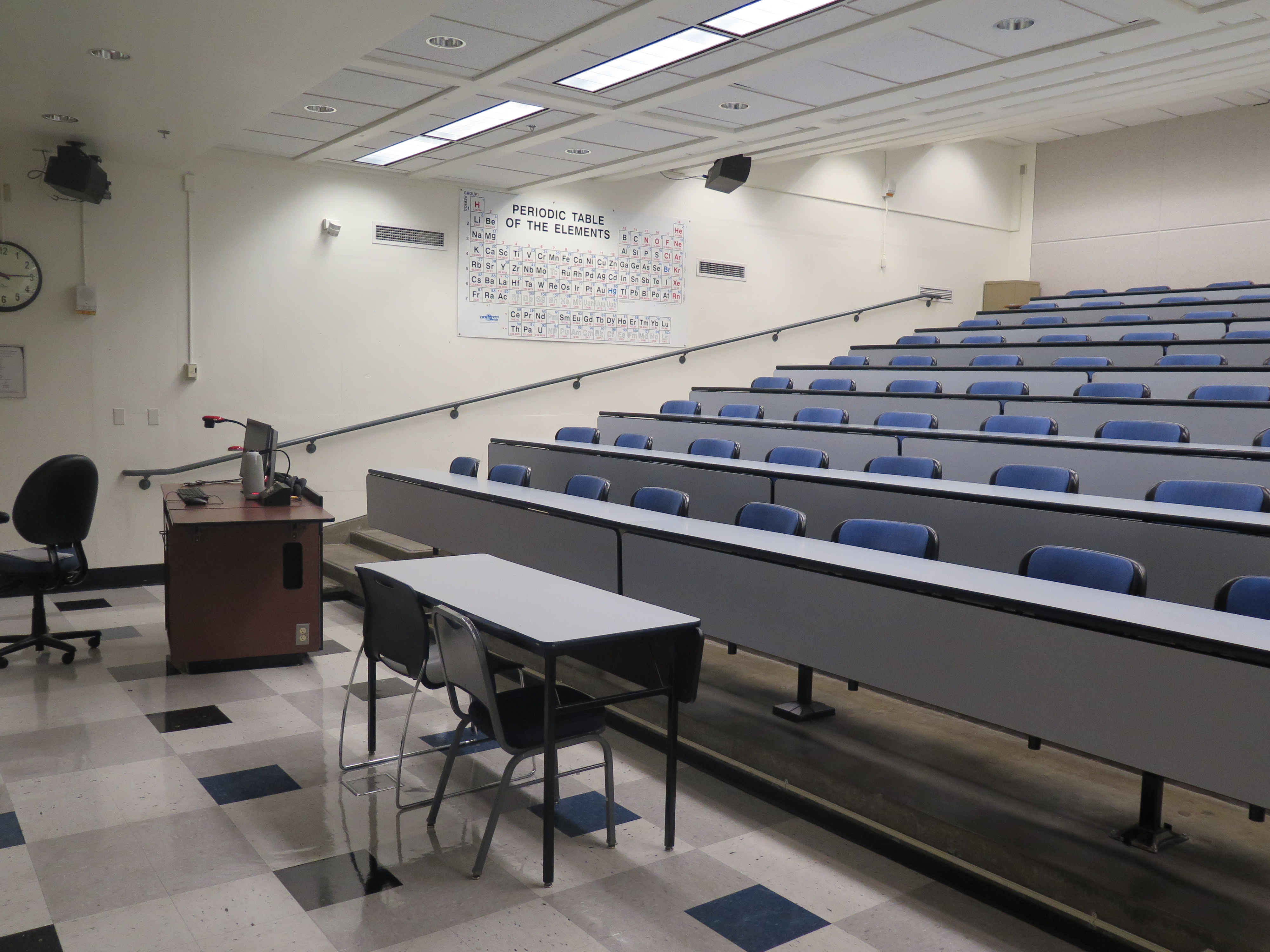 Room Consists of hard floors, Stationary rows of tables and chairs, a white board and podium are both located at the front of the room. this room also uses a Rear Screen projector, it is the large Gray surface next to the white board