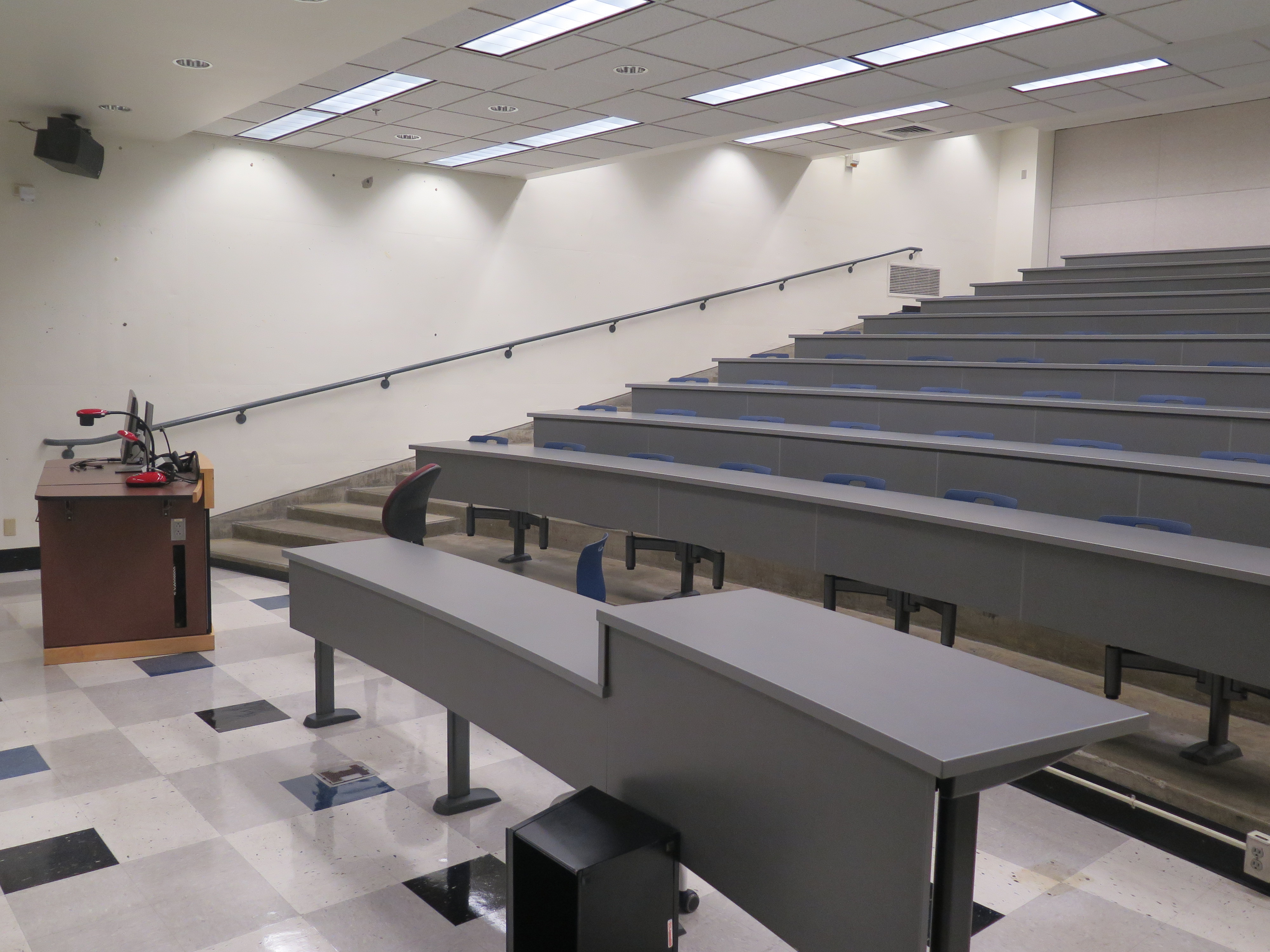 Room Consists of hard floors, Stationary rows of tables and chairs, a white board and podium are both located at the front of the room. this room also uses a Rear Screen projector, it is the large Gray surface next to the white board