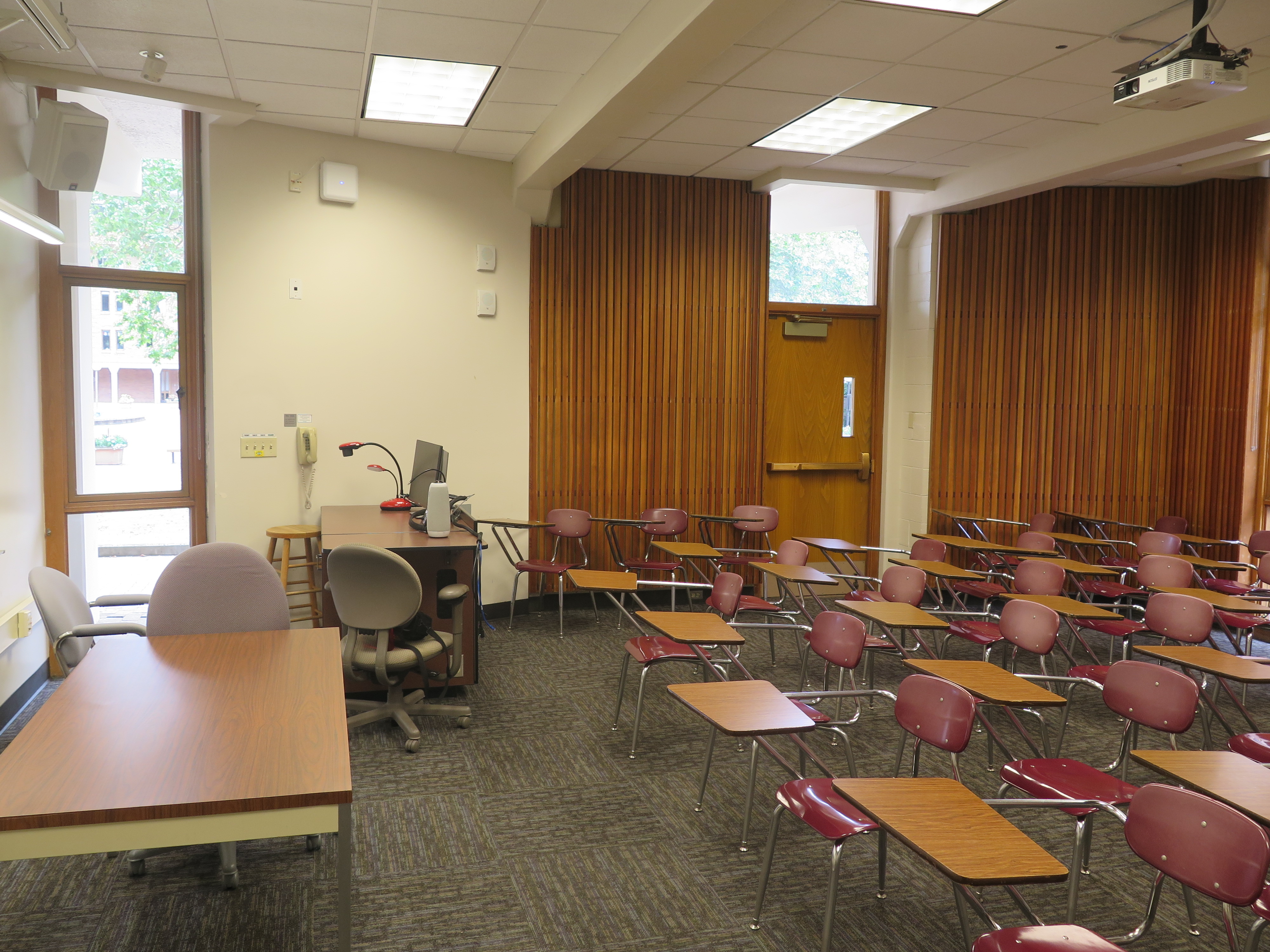 Room consists of carpet, moveable tablet armchairs and a table, podium and whiteboard located at the front of the room. 