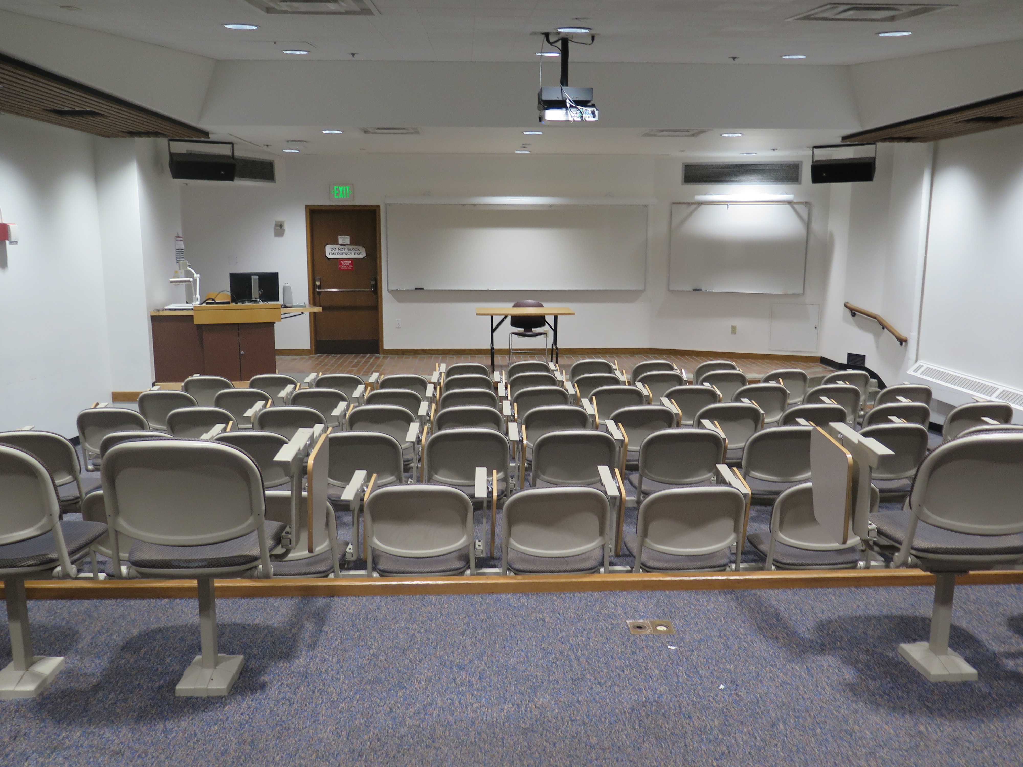Room Consists of carpet floors, Stationary rows of tablet armchairs, a stage, white board and podium are located at the front of the room. 